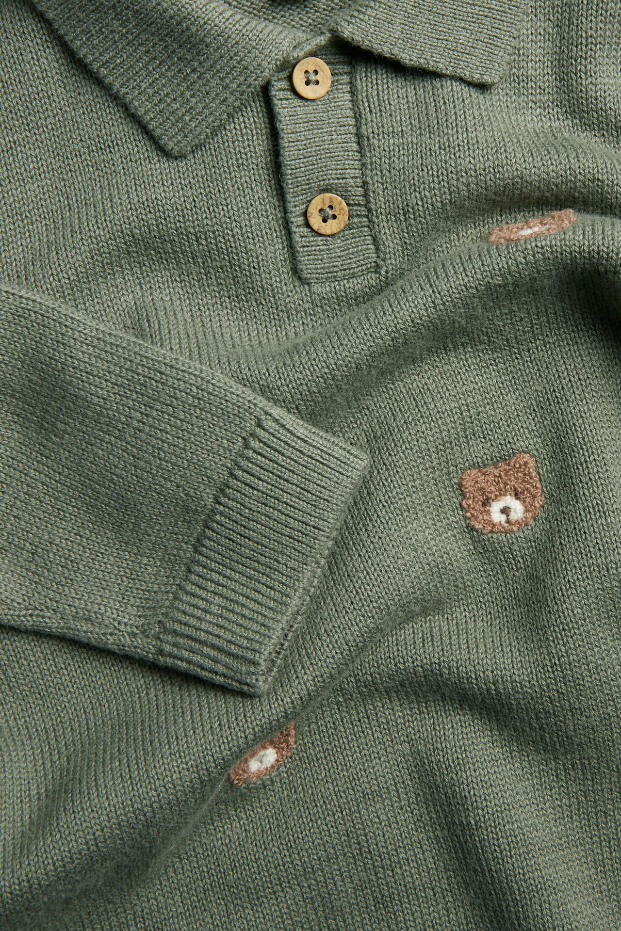 Nahaufnahme: Grüner Strickpullover mit Kragen, zwei Holzknöpfen, Rippenbündchen und gestickten Bären.