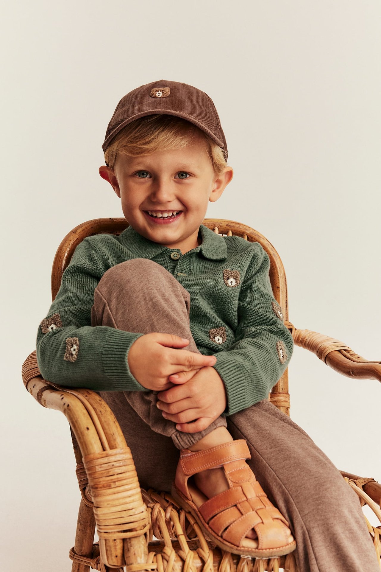 Vorderansicht: Kind in grünem Strick-Poloshirt mit Bärenstickerei, brauner Kappe, Jogginghose und Sandalen.