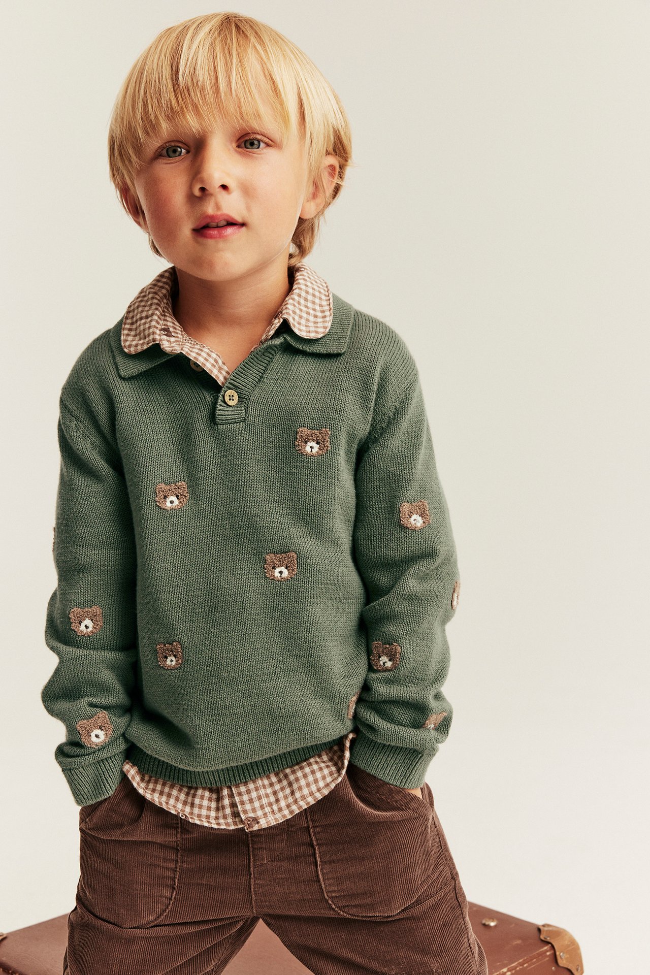 Kid in green knitted polo sweater with bear embroideries, brown checkered shirt, and brown corduroy pants.
