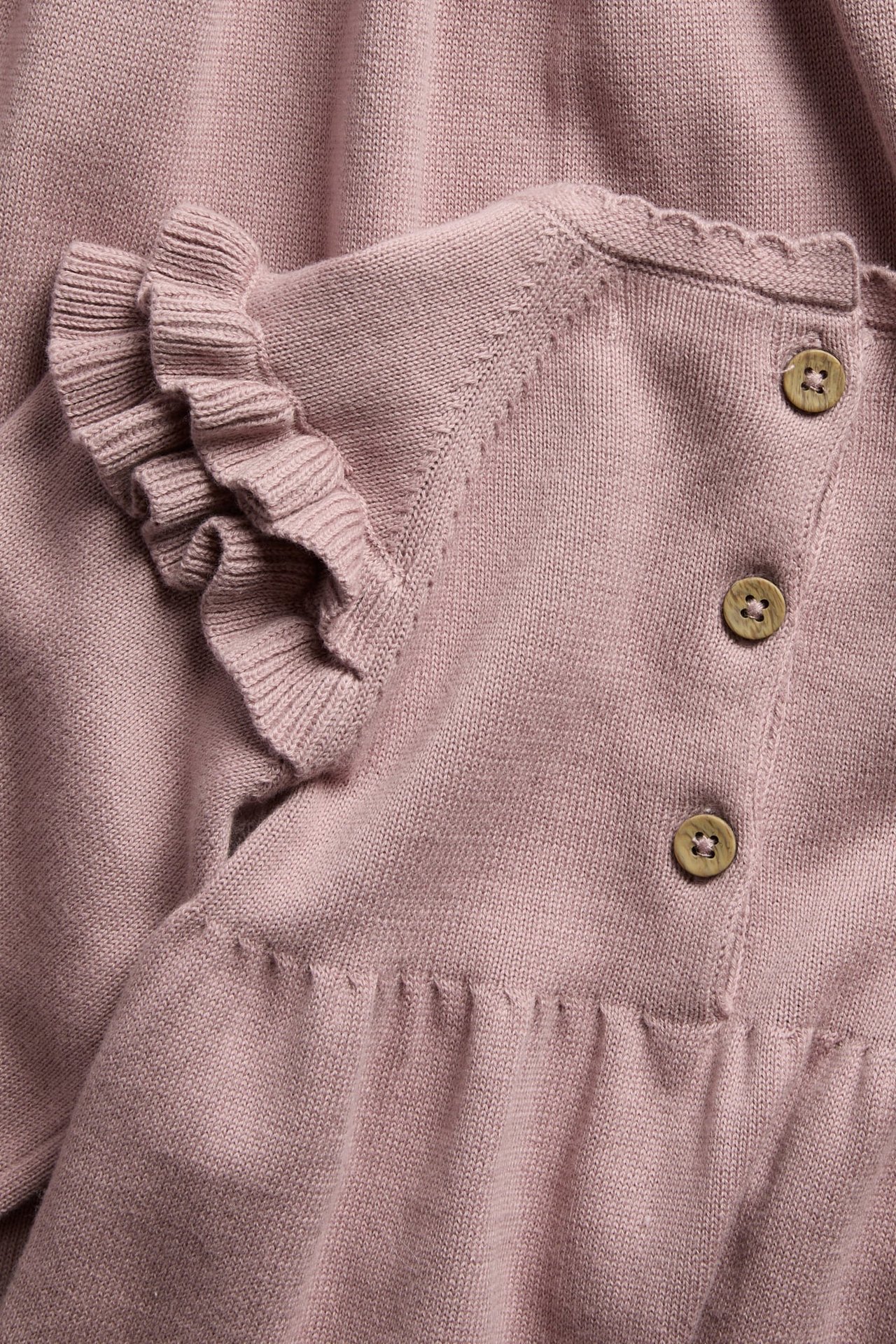 Nahaufnahme eines rosa gestrickten Babykleidungsstücks mit Rüschenärmeln, Holzknöpfen und gewelltem Saum.