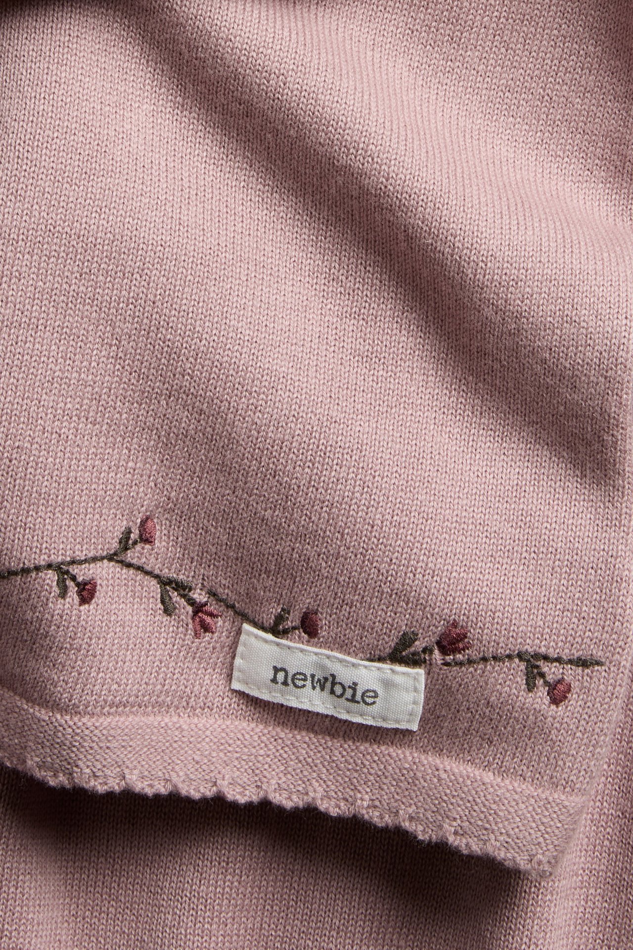 Nahaufnahme eines altrosafarbenen gestrickten Babykleidungsstücks mit besticktem Blumendetail und weißem Etikett.