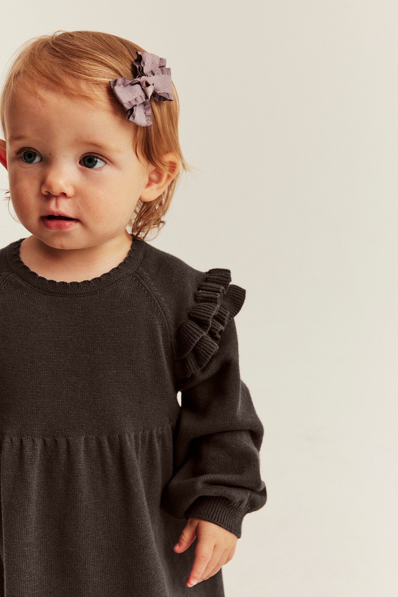 Close-up front view of a baby wearing a dark brown knitted ruffle-shoulder dress with a purple hair bow.