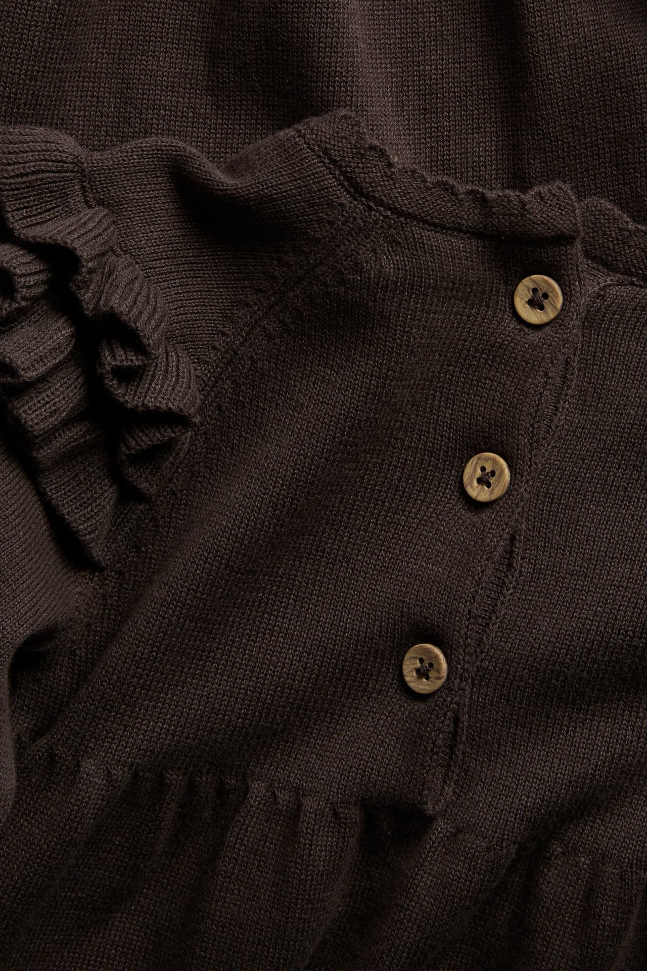 Nahaufnahme eines dunkelbraunen Strickoberteils mit Wellenkante, Rüschen und Holzknöpfen.