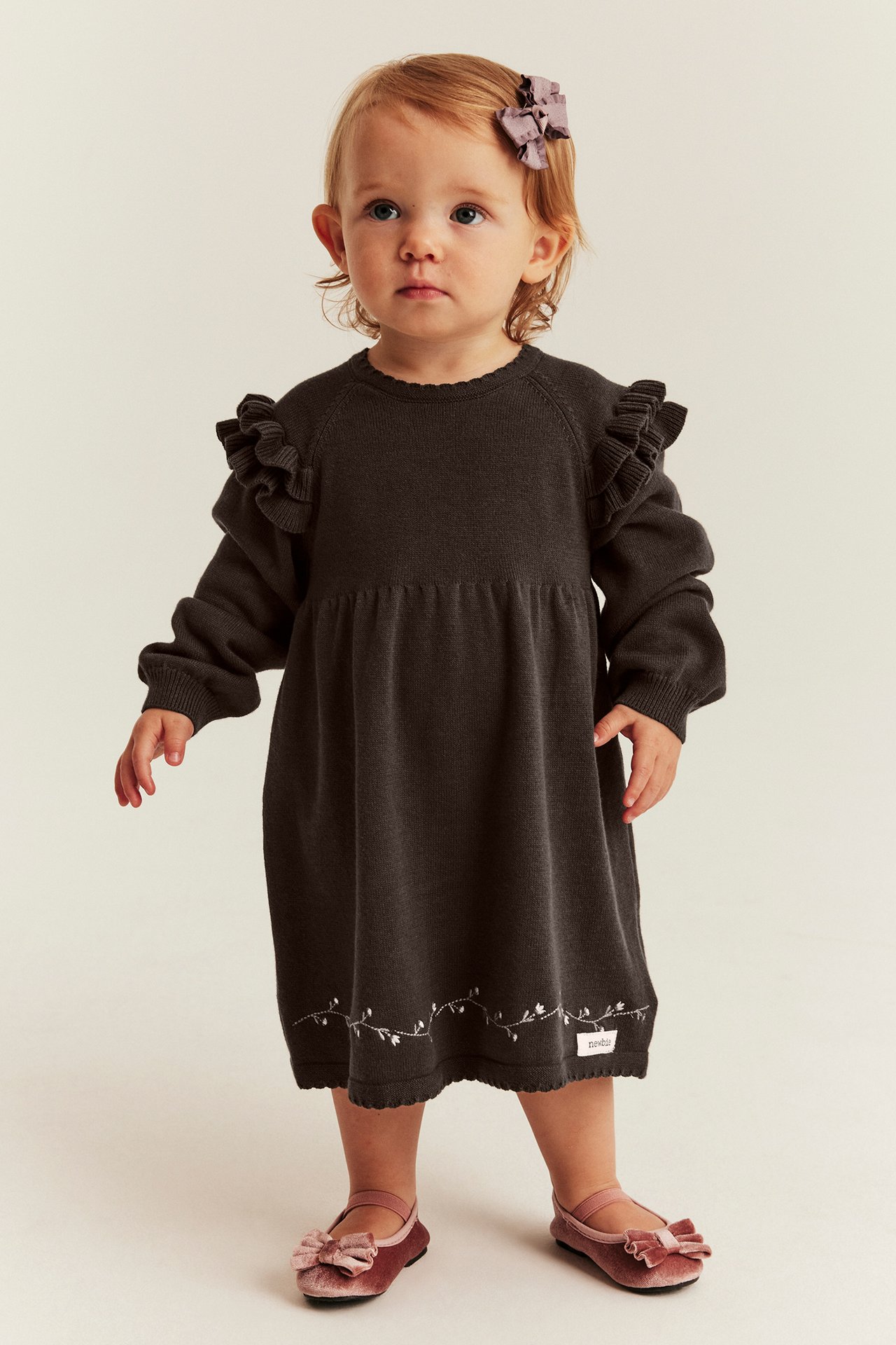 Baby in dunkelbraunem Strick-Rüschenkleid, rosa Samt-Ballerinas und lila Schleifen-Haarspange.