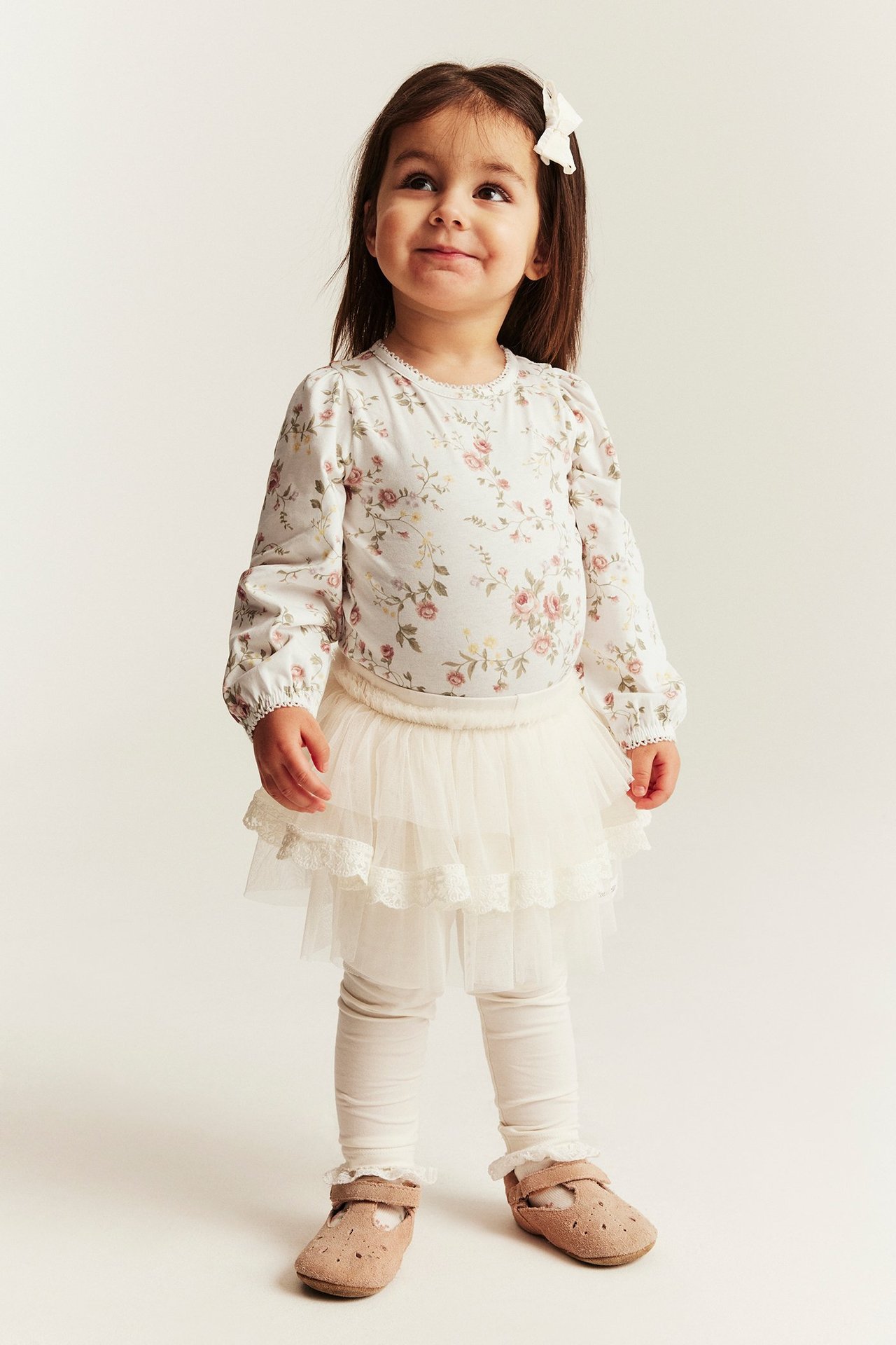 Front view: Kid wears an off-white floral long-sleeved bodysuit, cream tulle skirt, off-white leggings, tan shoes, white bow.