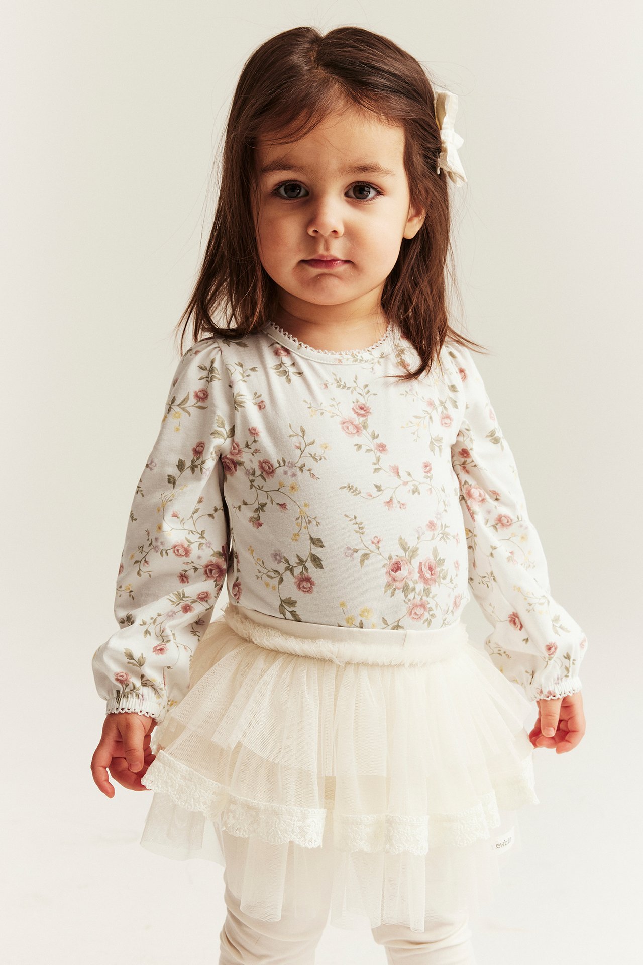 Front view: Kid in off-white floral long-sleeved bodysuit, tiered tulle skirt, leggings, and a hair bow.