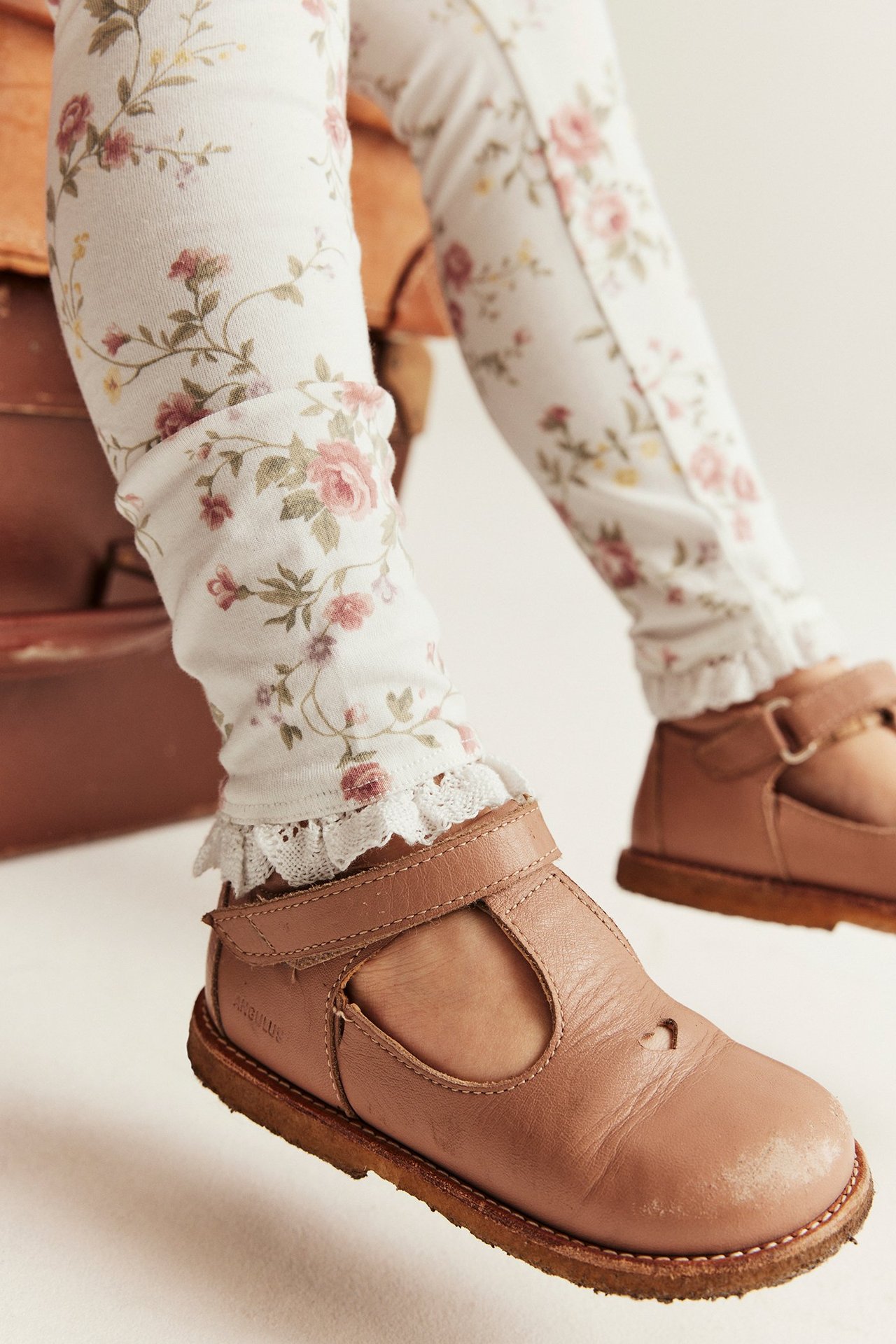 Close-up of a kid's legs wearing off-white floral leggings with lace ruffles, paired with brown T-strap shoes.