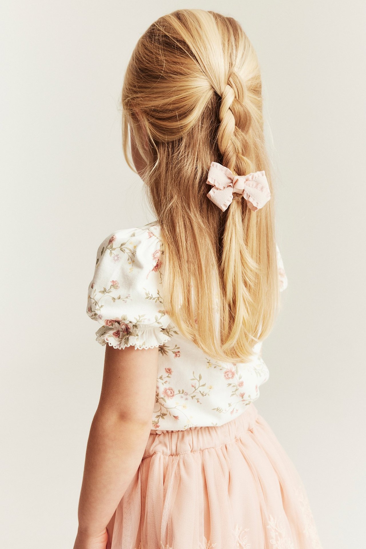 Rückenansicht: Kind trägt off-weißes geblümtes Jersey-Puffärmel-Top, einen rosa Tüllrock und eine rosa Haarschleife.