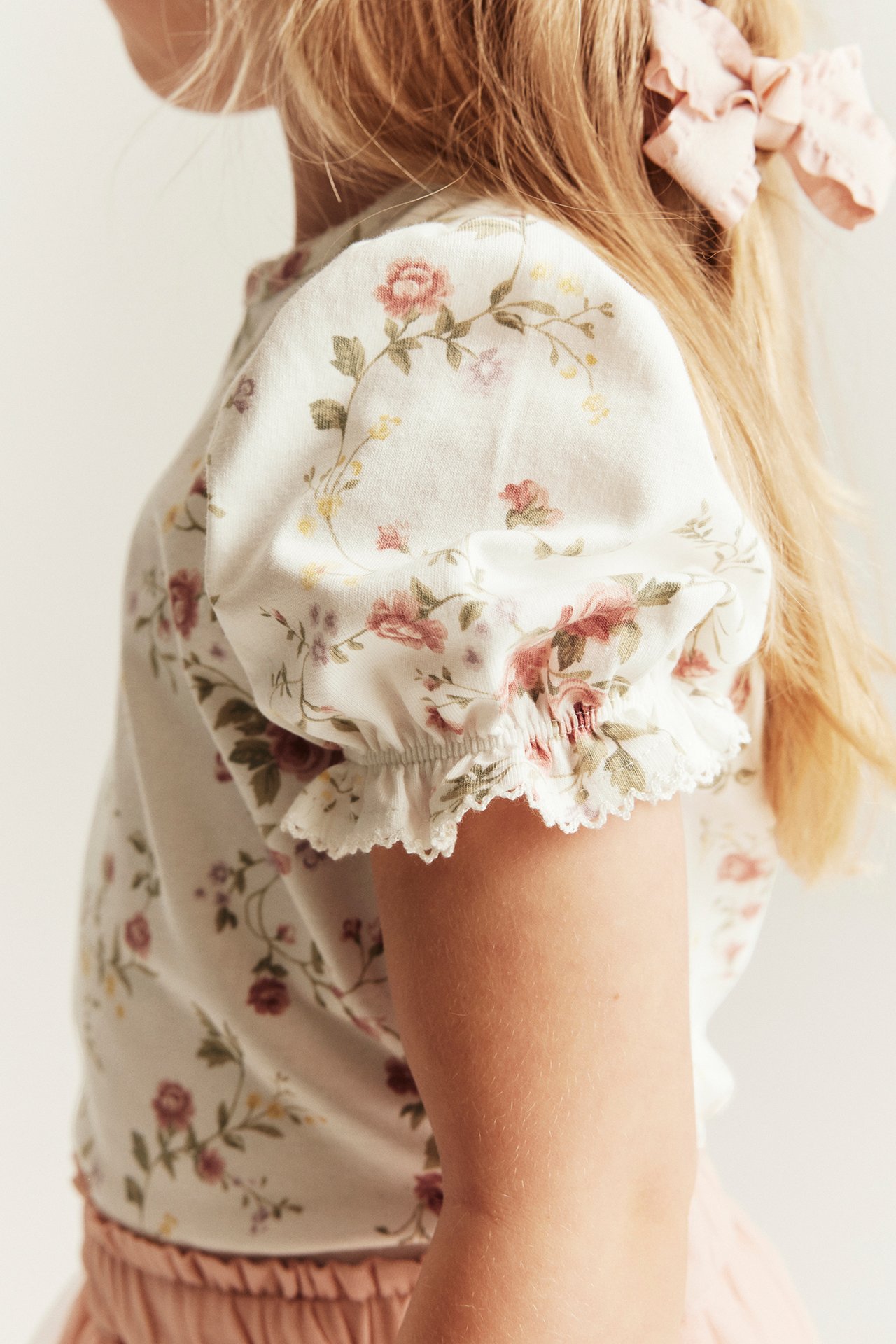 Seitenansicht: Kind trägt cremeweißes geblümtes Jersey-Top mit Puffärmeln, rosa Rock, rosa Haarschleife.