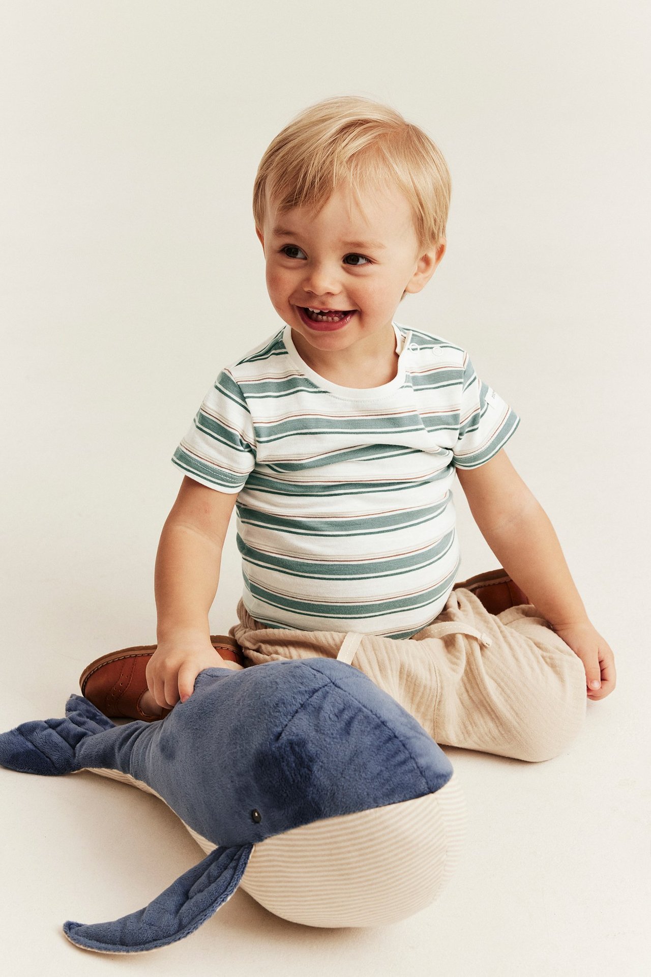 Lachendes Baby in gestreiftem, cremefarbenem Baumwollbody und beiger Hose, hält einen Blauwal.