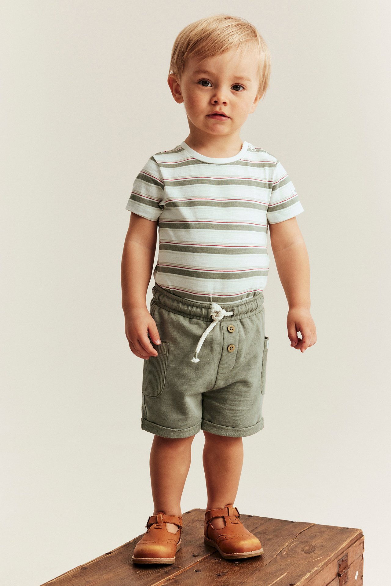 Front view of a baby wearing an off-white striped short-sleeved cotton body, green shorts, and brown sandals.