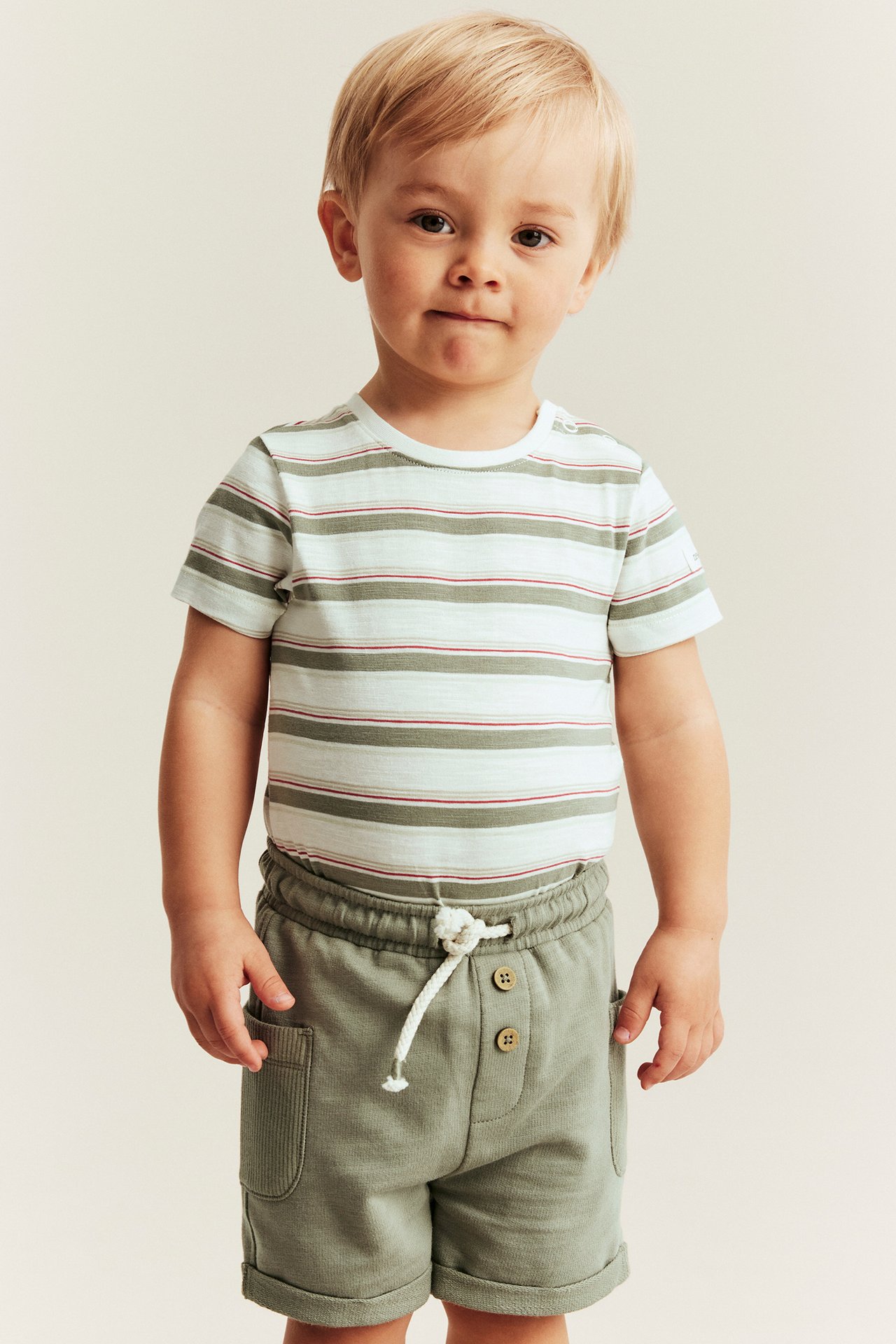 Front view of a baby boy wearing an off-white striped short-sleeved bodysuit and olive green shorts.