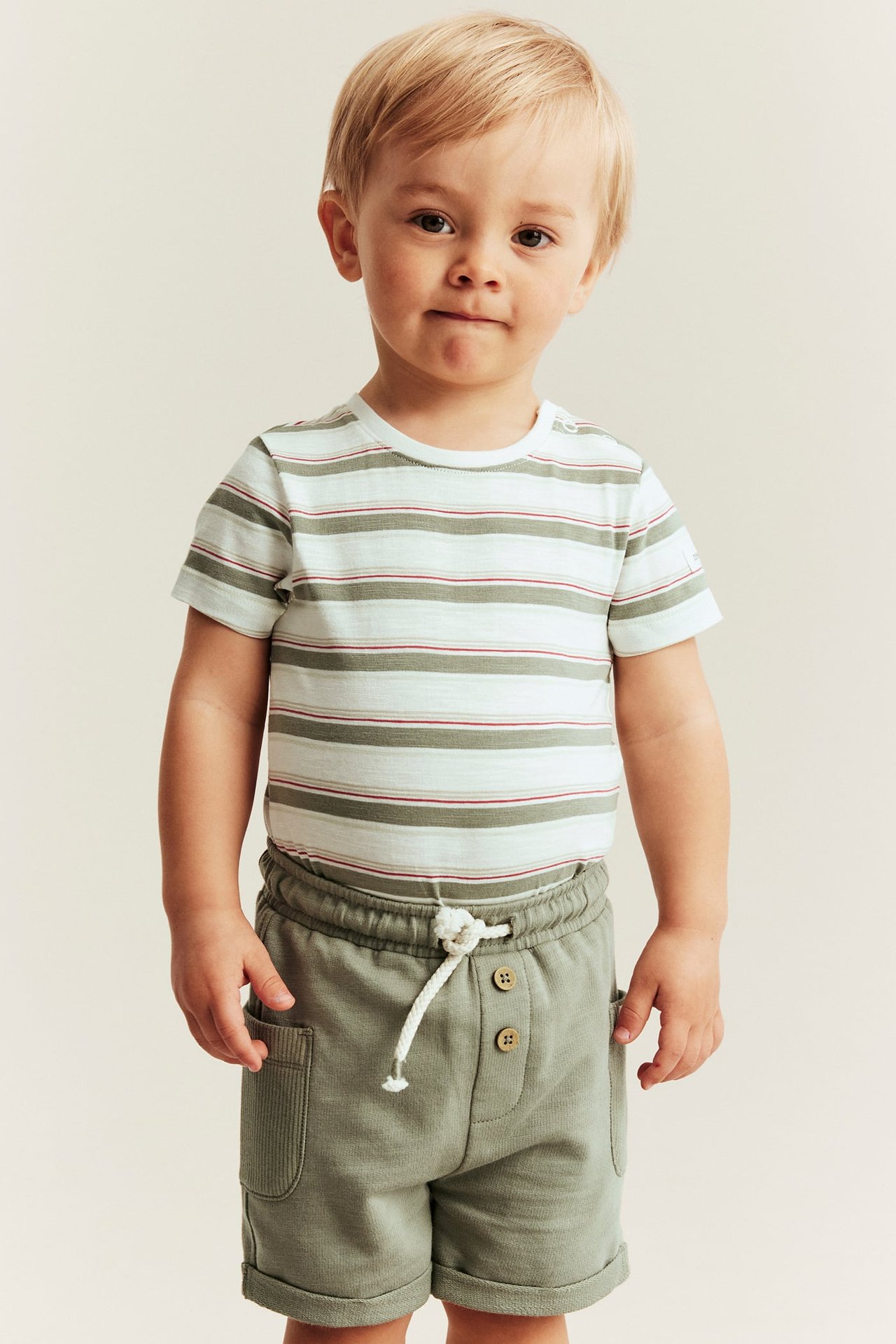 Front view of a baby boy wearing an off-white striped short-sleeved bodysuit and olive green shorts.