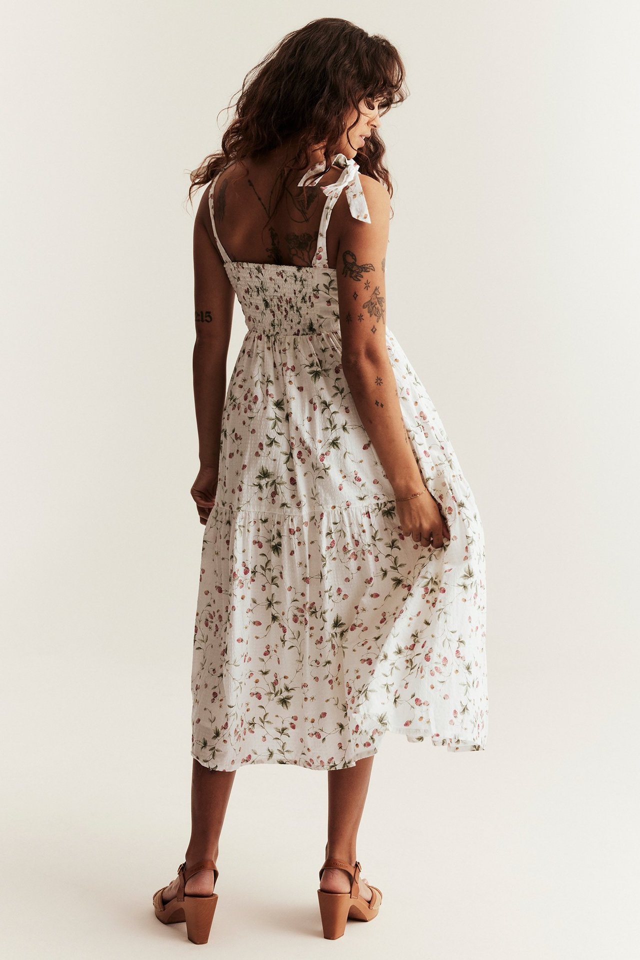 Back view of a woman in an offwhite smocked tiered cotton dress with strawberry print and brown heeled sandals.