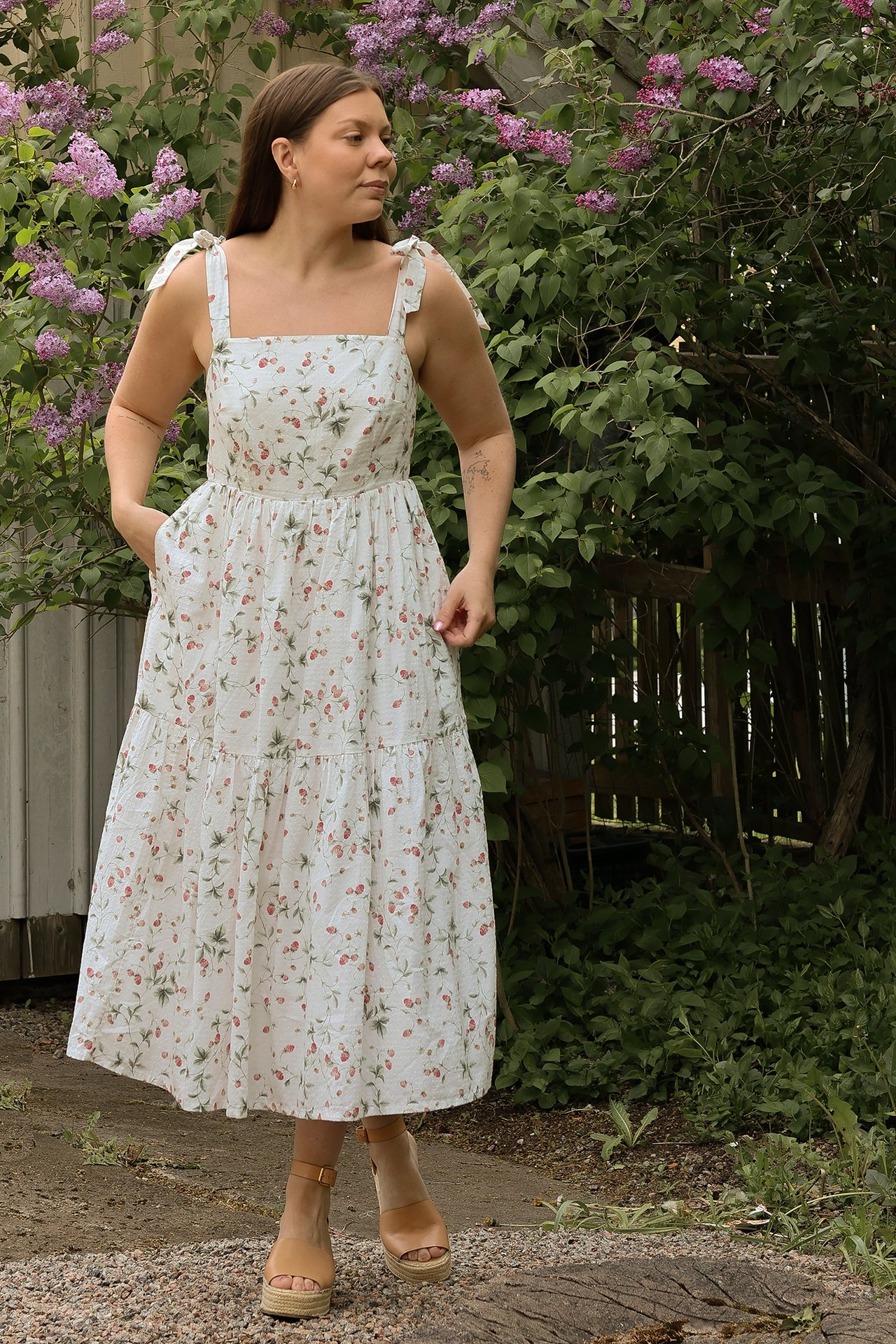 Vorderansicht: Frau in einem cremeweißen Baumwollkleid mit Erdbeermuster, Stufenrock, Bindeträgern und beigefarbenen Keilsandalen.
