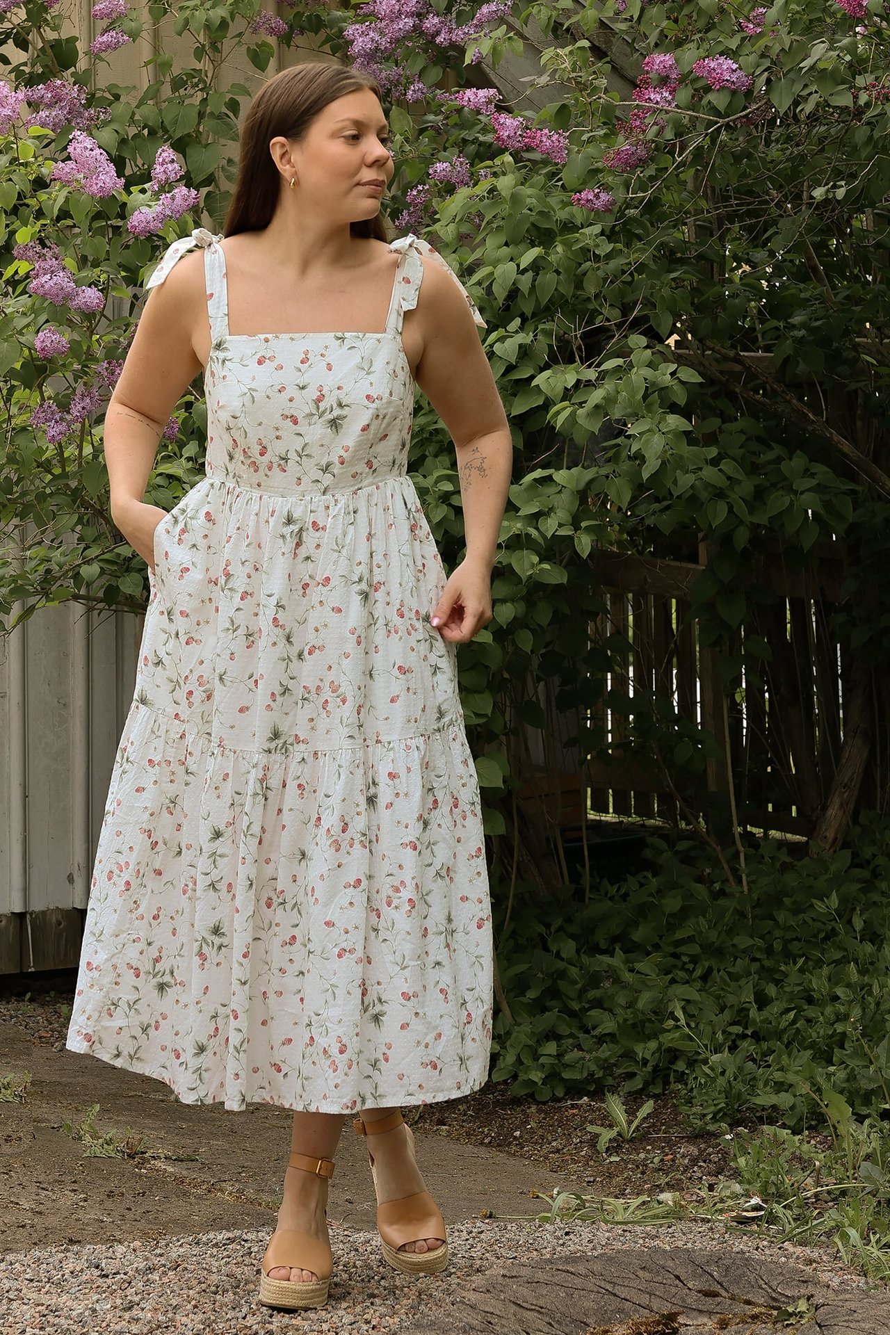 Front view: woman in an offwhite strawberry print tiered cotton dress with tie straps, pockets, and tan wedge sandals.
