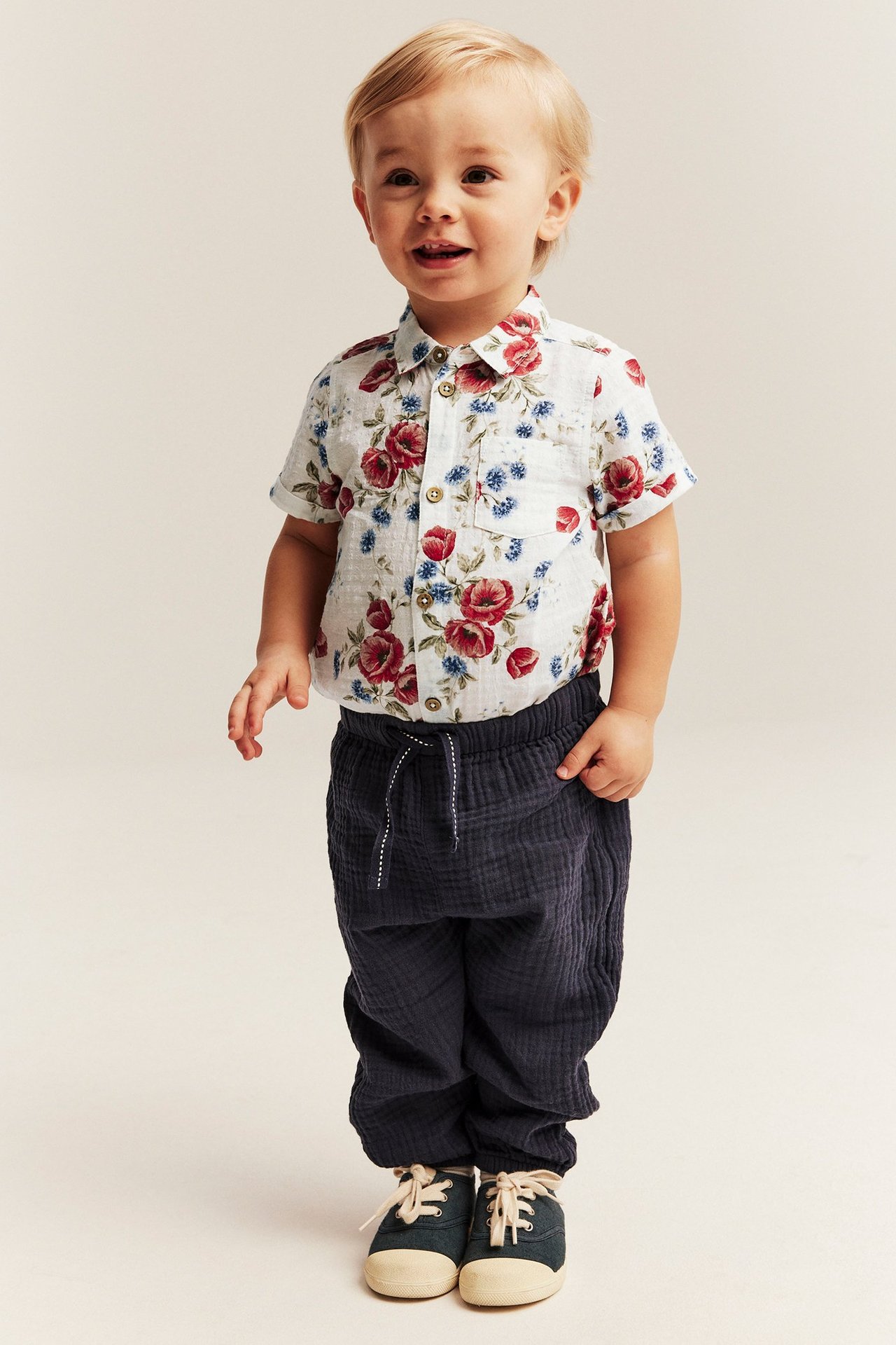 Vorderansicht eines Baby-Jungen in einem cremeweißen geblümten Baumwoll-Kurzarm-Shirtbody, dunkelblauer Hose und navyfarbenen Schuhen.