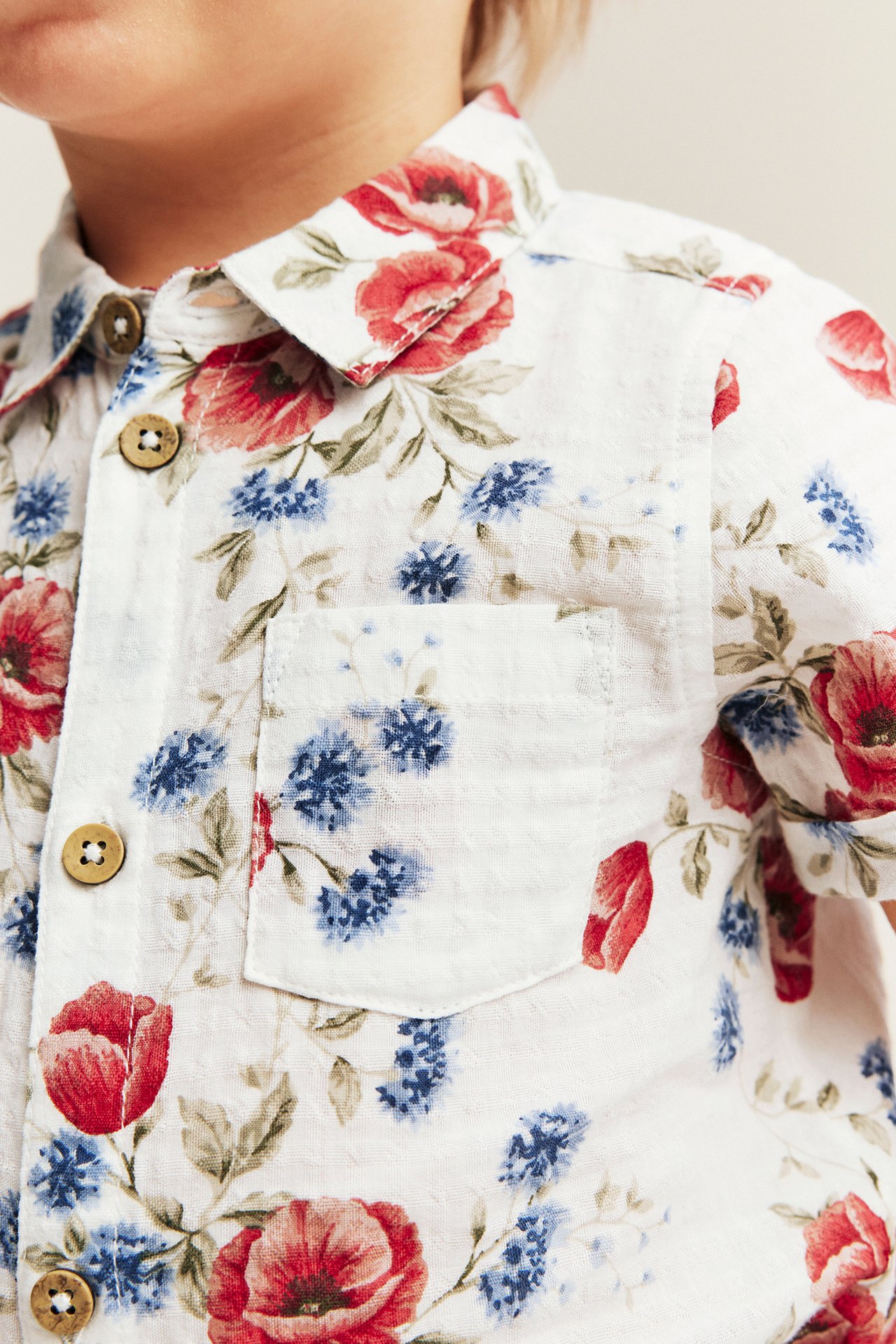 Nahaufnahme: Kind in cremeweißem, geblümtem Kurzarm-Shirtbody mit roten, blauen Blumen, Knopfleiste und Tasche.
