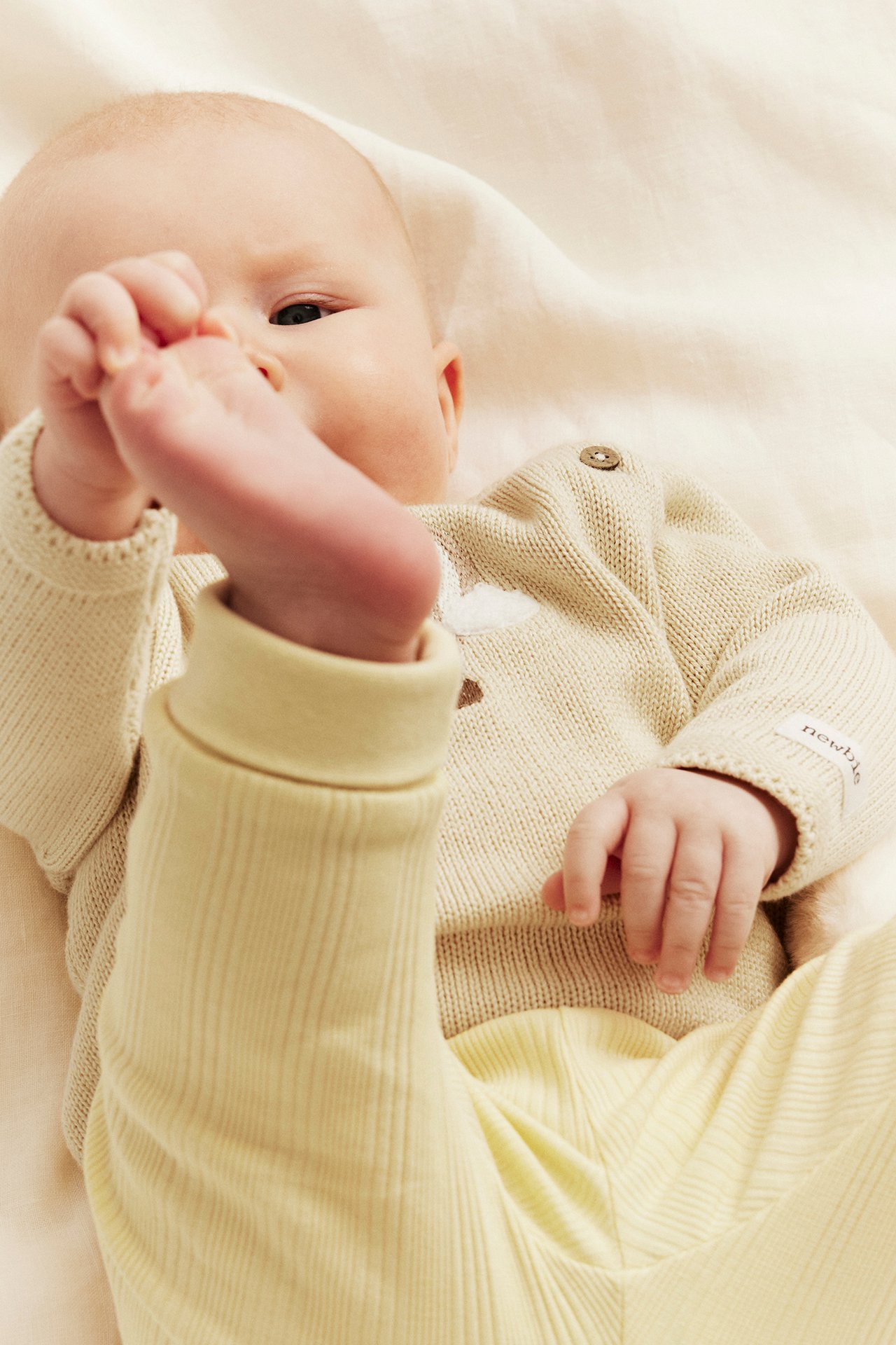 Baby in cremefarbener feingestrickter Baumwollstrickjacke mit Heißluftballon-Stickerei und hellgelber Rippenhose.