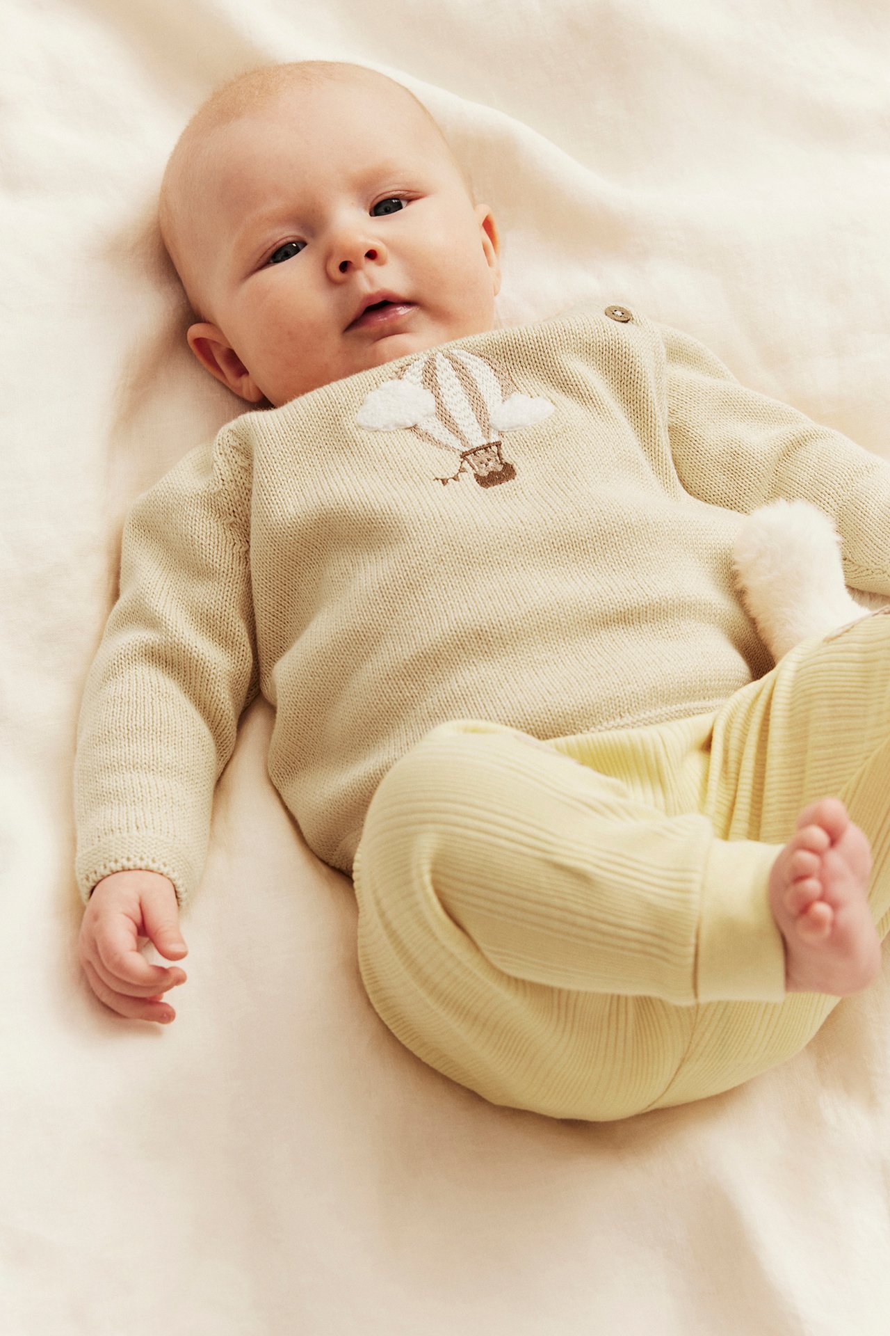 Front view: Baby in a light yellow fine-knit cotton cardigan with hot air balloon embroidery and matching ribbed pants.