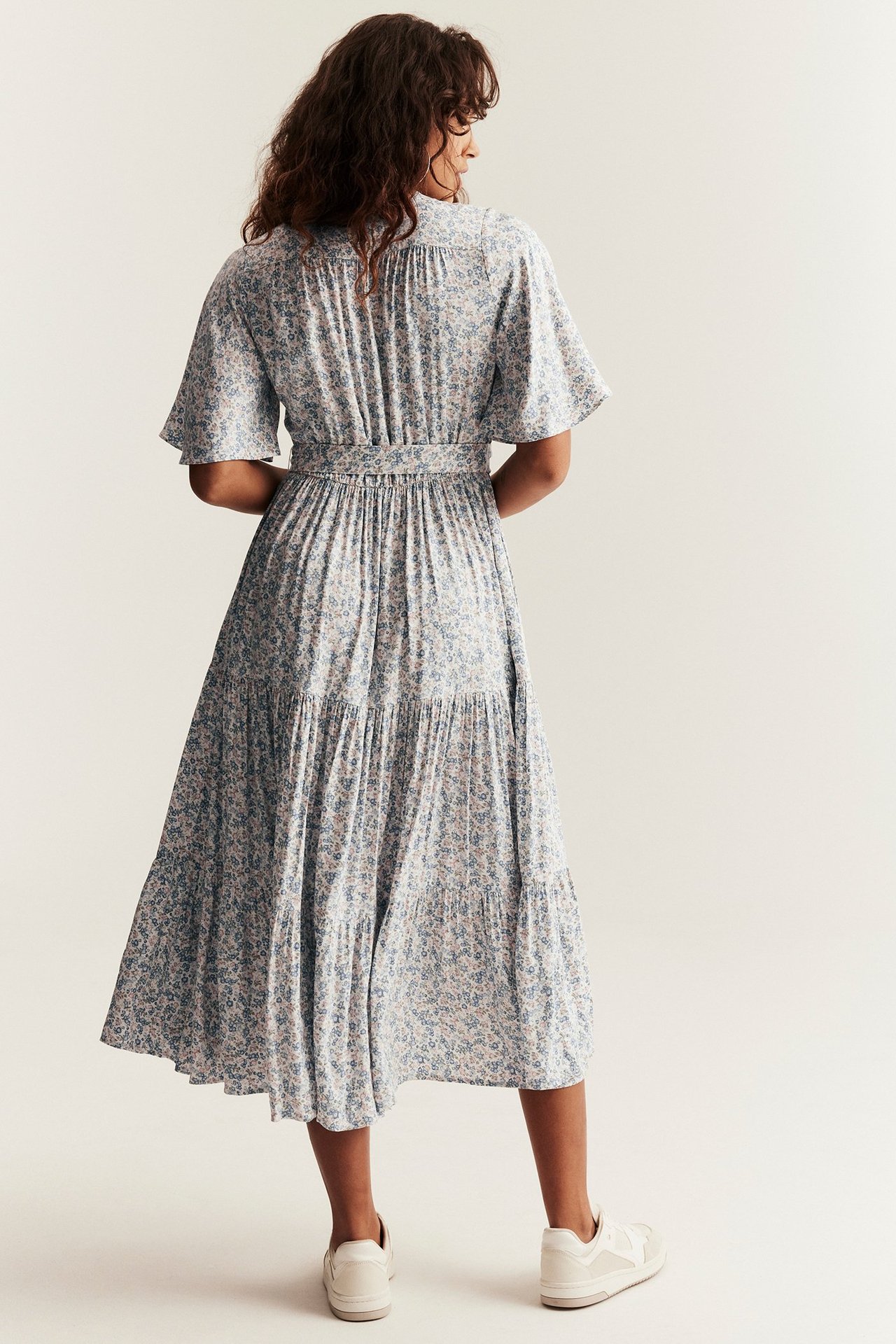 Back view: Woman in off-white floral viscose midi dress with short sleeves, tied belt. White sneakers.