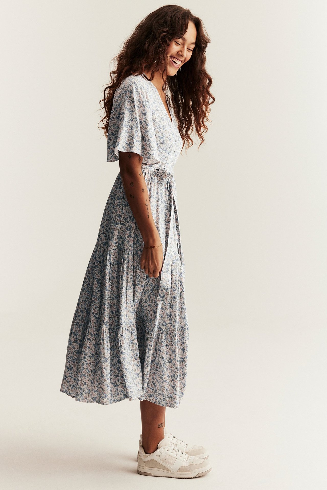 Seitenansicht einer Frau in gebrochen weißem Blumen-Viskose-Midikleid mit kurzen Ärmeln, gebundenem Gürtel und weißen Sneakern.