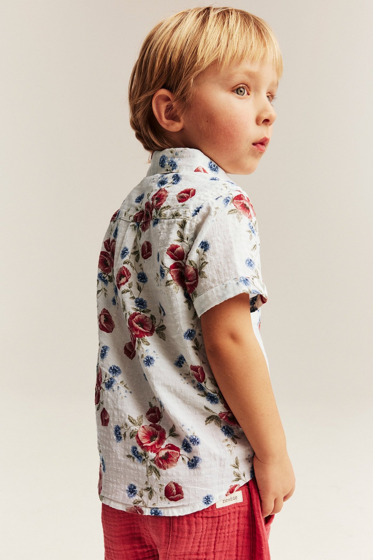 Side view of a kid in an off-white cotton short-sleeved floral shirt and red textured shorts.
