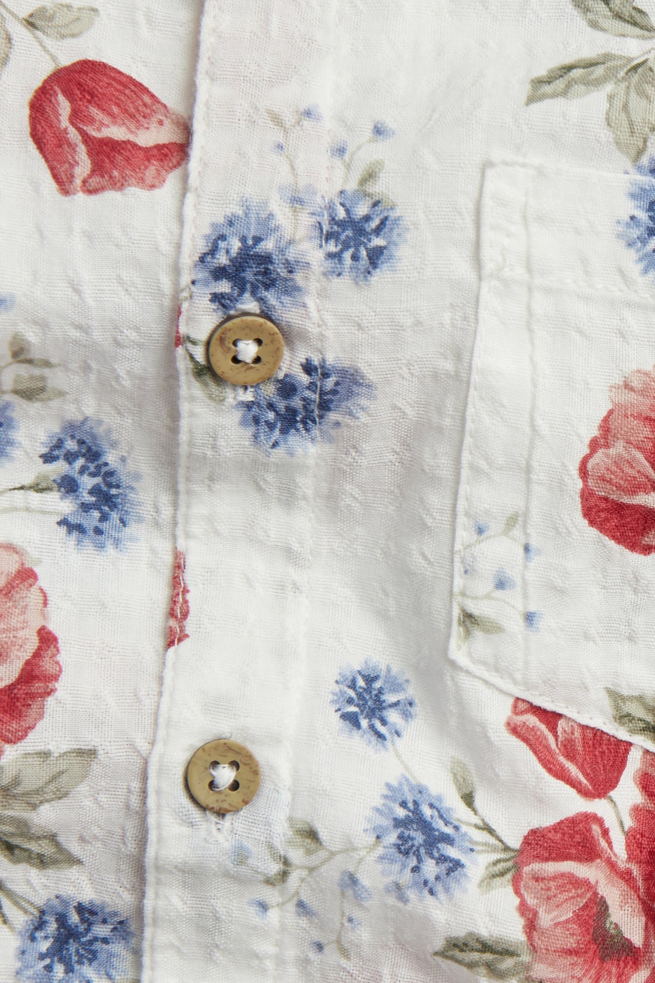 Nahaufnahme: cremefarbenes kurzärmeliges Baumwollhemd mit rot-blauem Blumenmuster, Brusttasche und braunen Knöpfen.