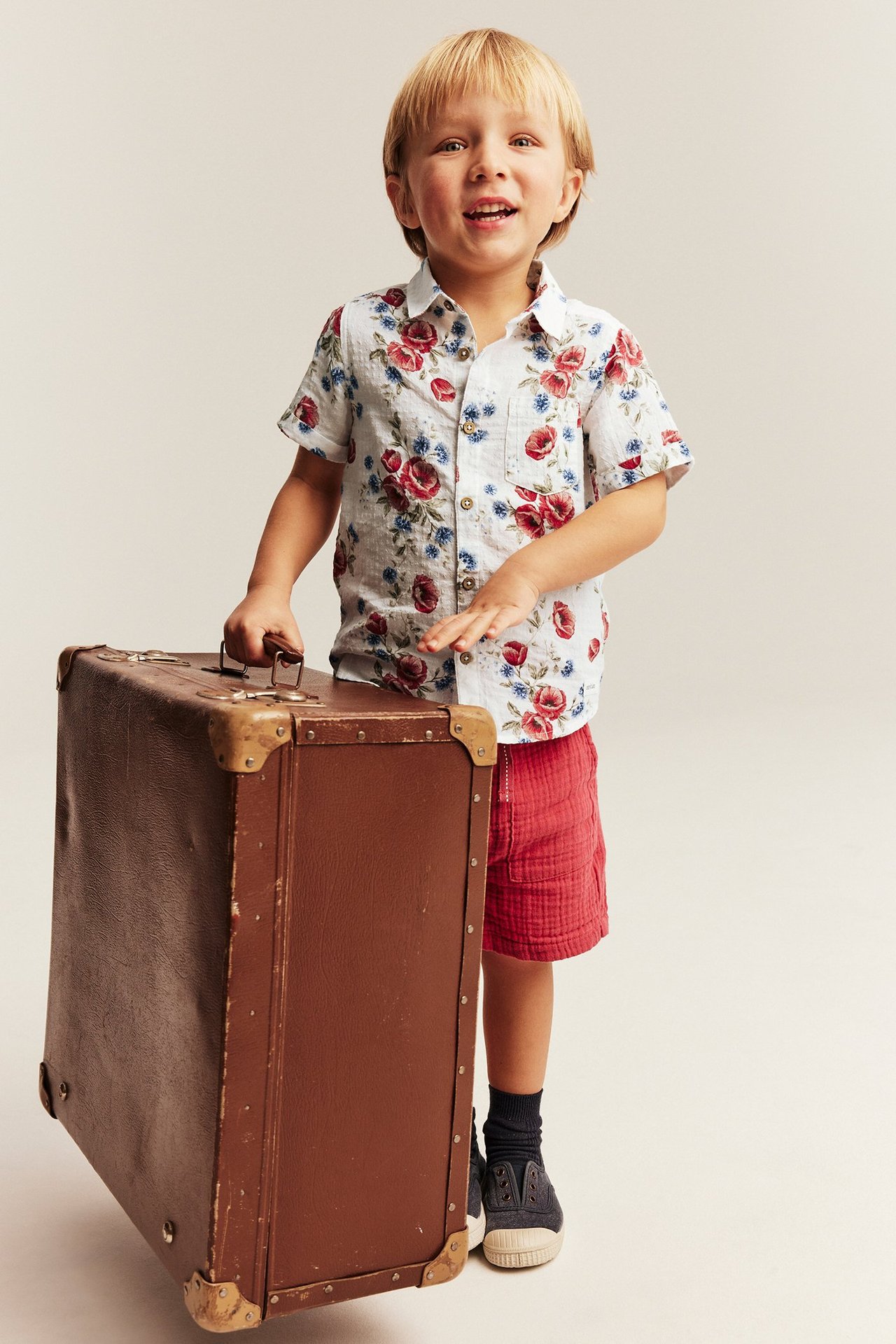 Vorderansicht: Kind in offwhite Kurzarm-Blumenhemd, roten Shorts, hält einen braunen Koffer.