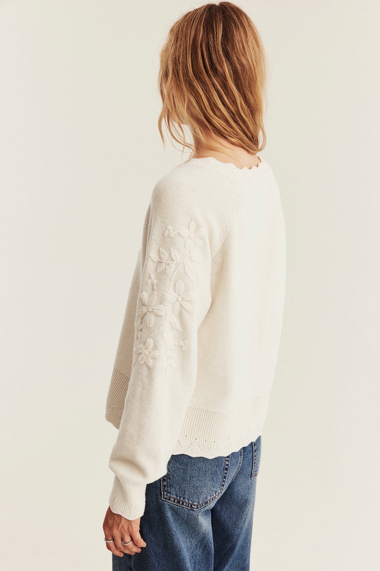 Back-left side view of a female wearing a cream embroidered long-sleeve sweater with scalloped details and blue jeans.