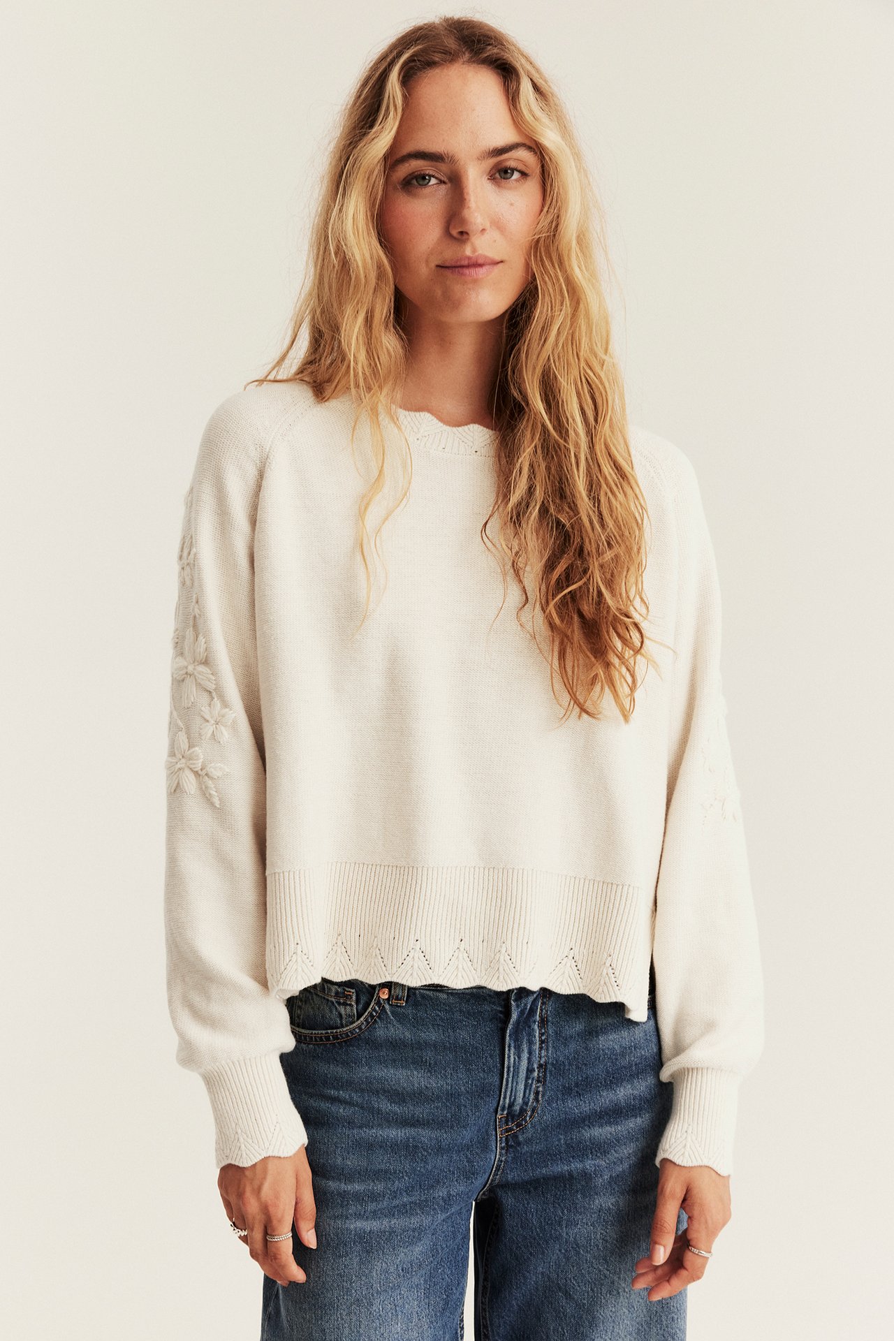 Front view of female model in off-white scallop-trim sweater with embroidered flowers and blue jeans.