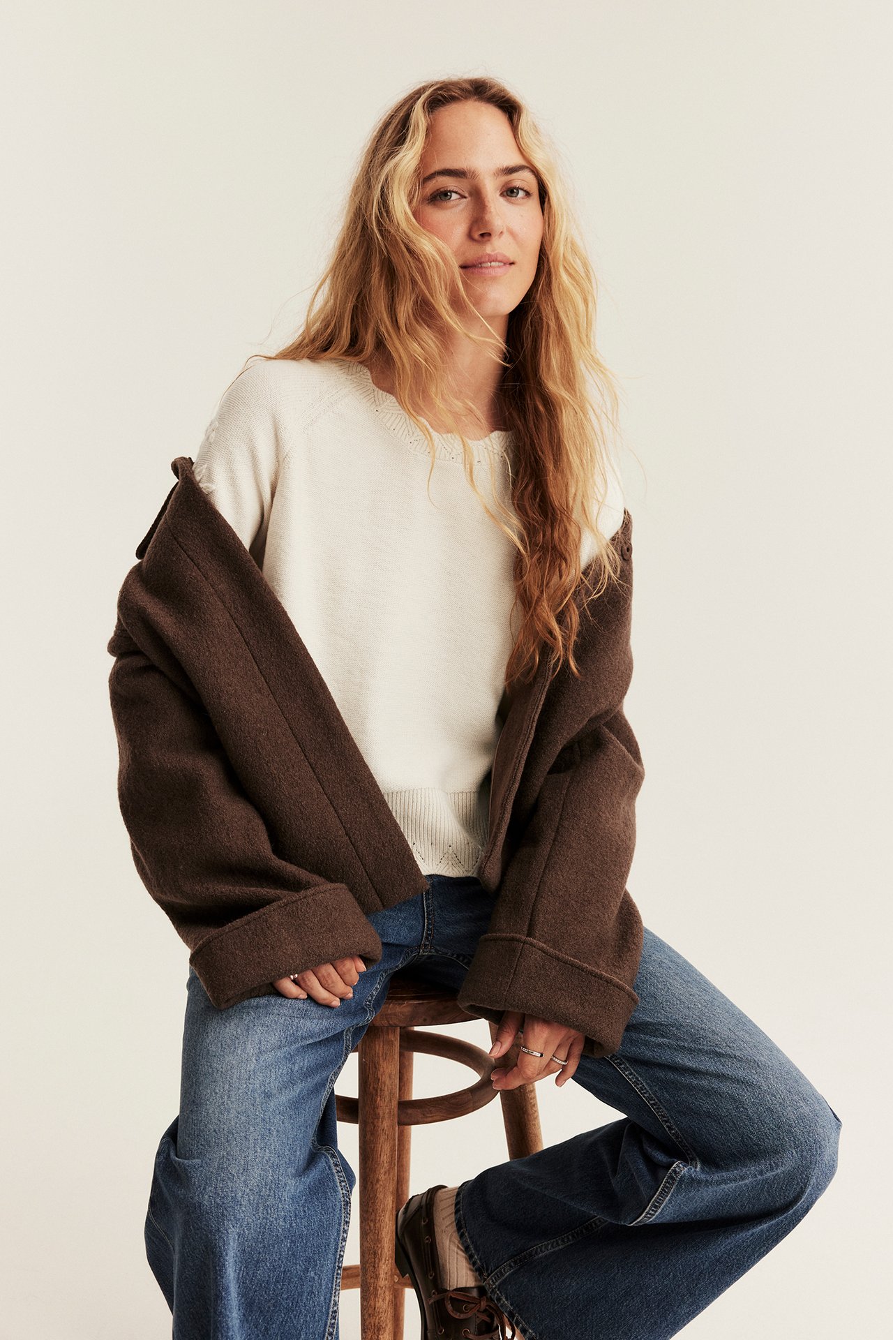 Front view of a woman wearing a brown relaxed fit jacket over a cream knit sweater and blue jeans.