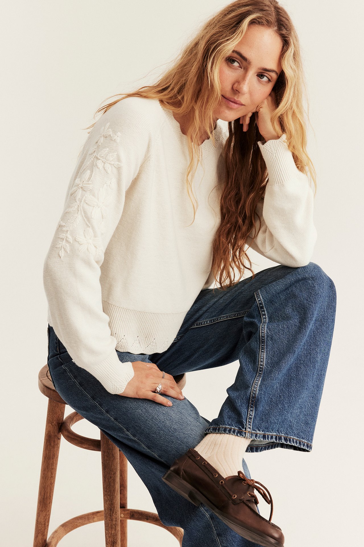 Front view, female model in off-white embroidered sweater, blue jeans, cream socks, brown boat shoes.