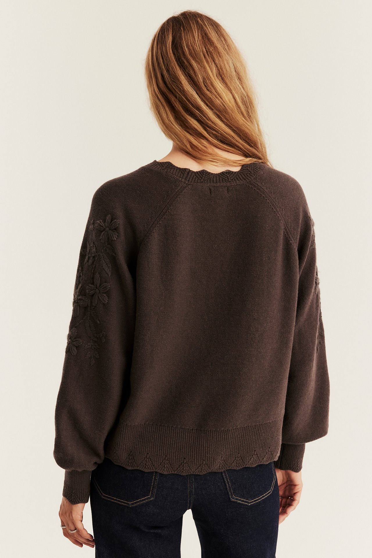 Back view of a woman in a dark brown knit sweater with floral embroidery and dark jeans.
