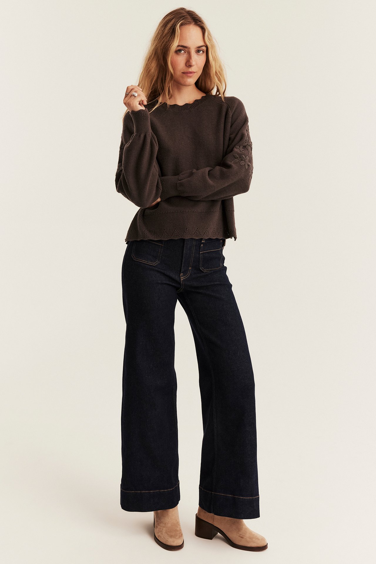 Dunkelbrauner Pullover mit Wellensaum und bestickten Ärmeln, weite Jeans und hellbraune Mules.