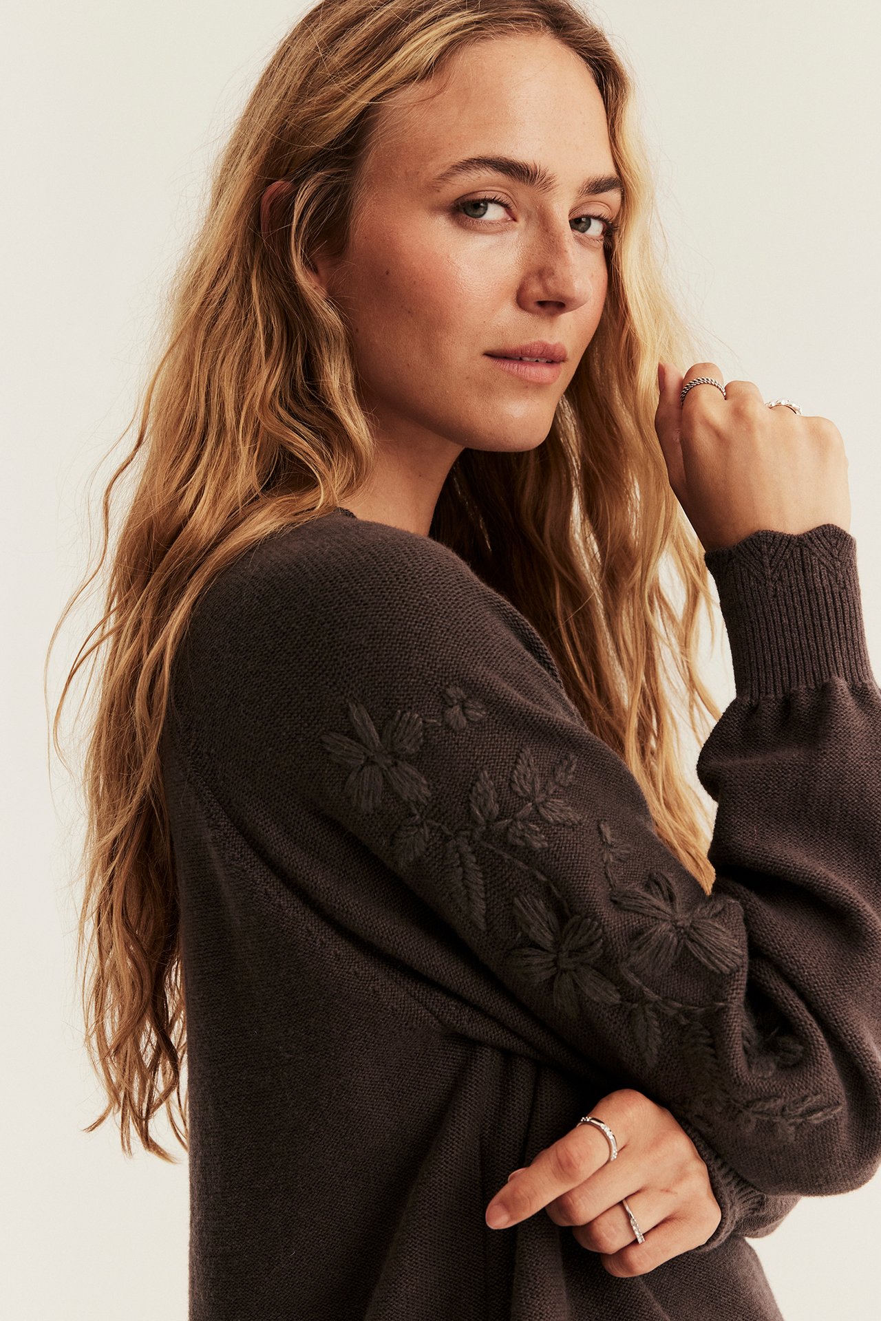 Nahaufnahme einer Frau in dunkelbraunem besticktem Pullover und Silberringen.