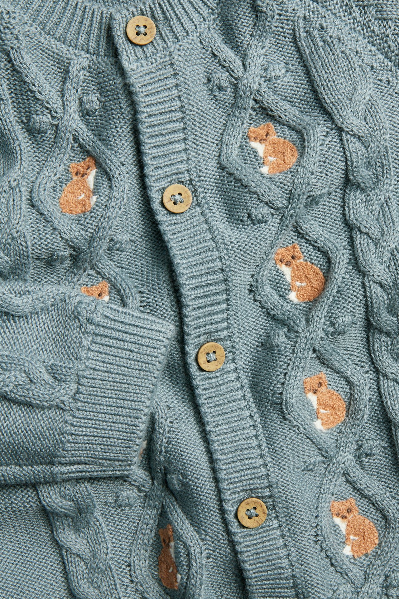 Nahaufnahme einer blauen Zopfmuster-Babyjacke mit Fuchsstickerei und Holzknöpfen.