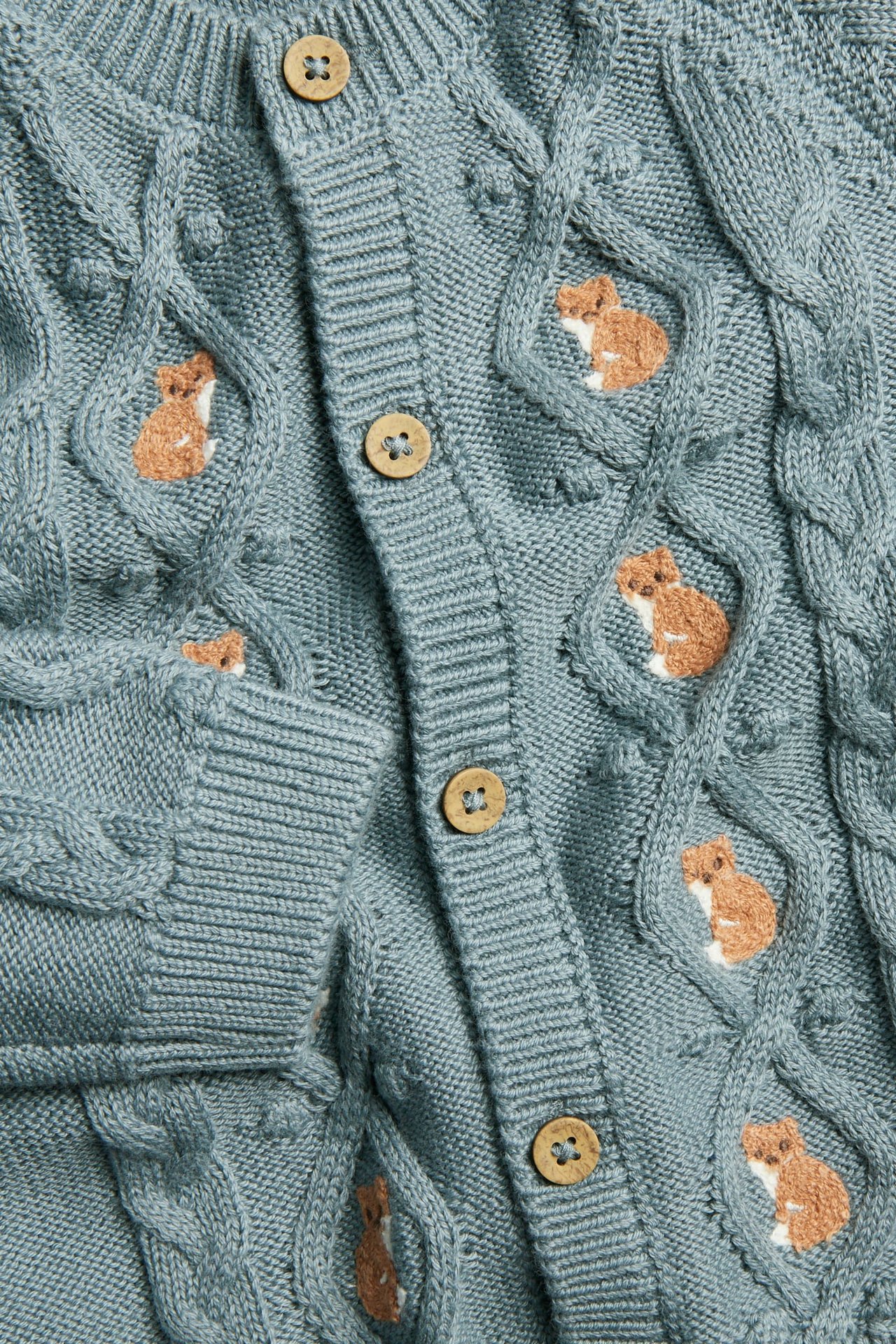 Nahaufnahme einer blauen Zopfmuster-Babyjacke mit Fuchsstickerei und Holzknöpfen.