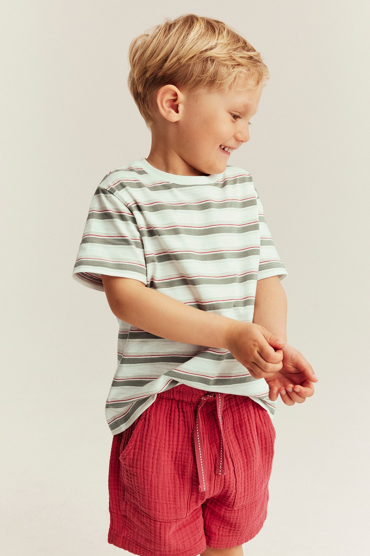 Front view of a kid in an off-white, striped cotton t-shirt with turned-up cuffs, and red shorts.