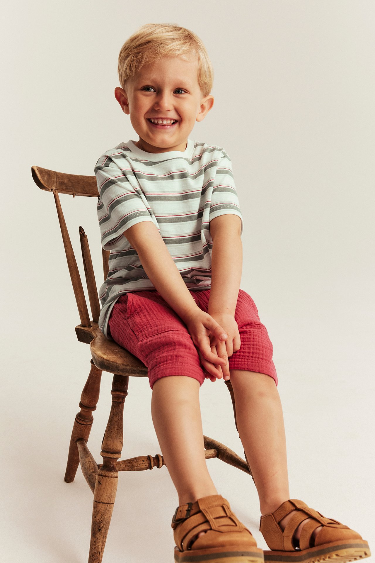 Front view: a smiling kid wears an off-white striped cotton t-shirt, red shorts, and brown sandals.