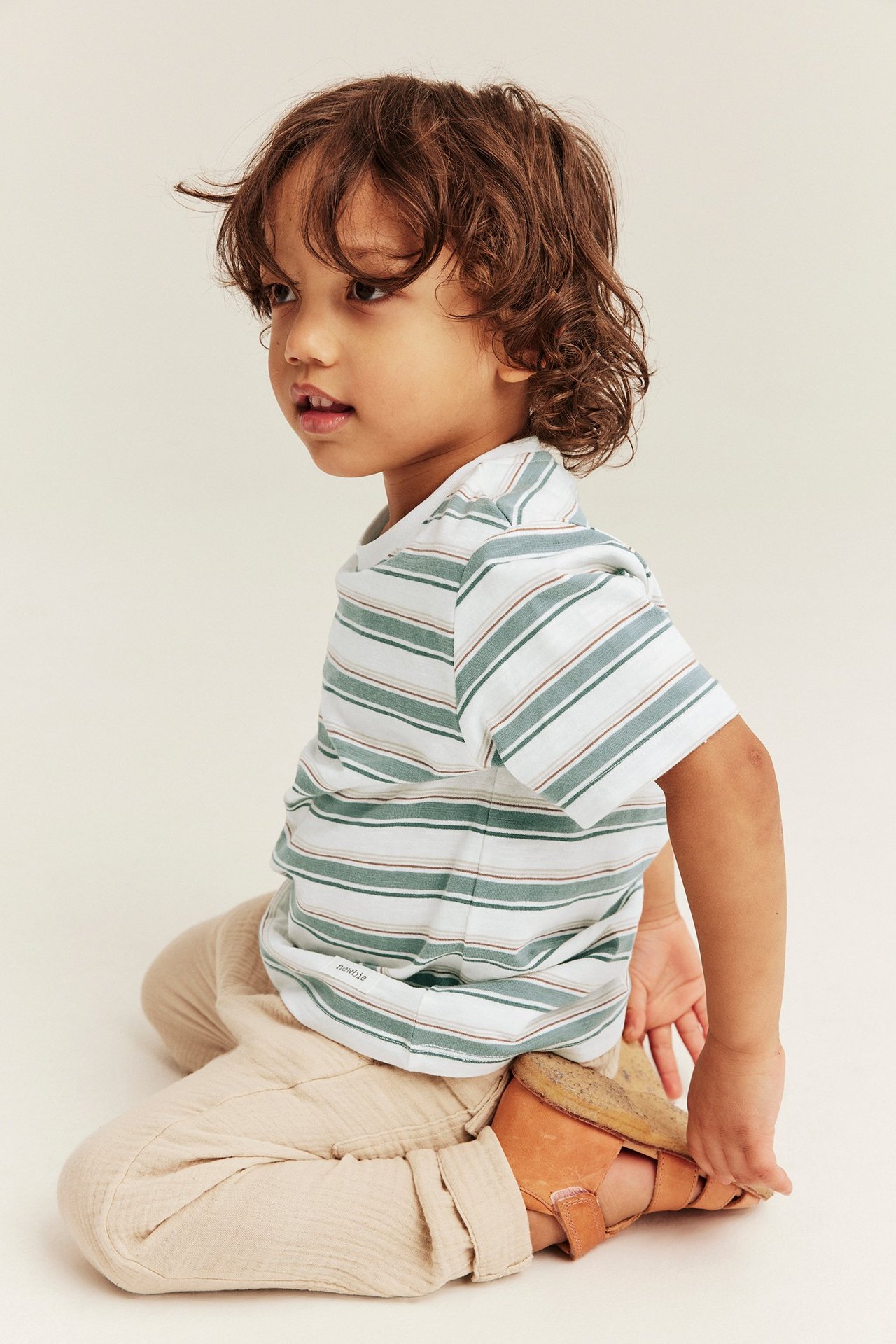 Seitenansicht eines Kindes: gestreiftes kurzärmeliges T-Shirt, beige Hose, braune Ledersandalen.