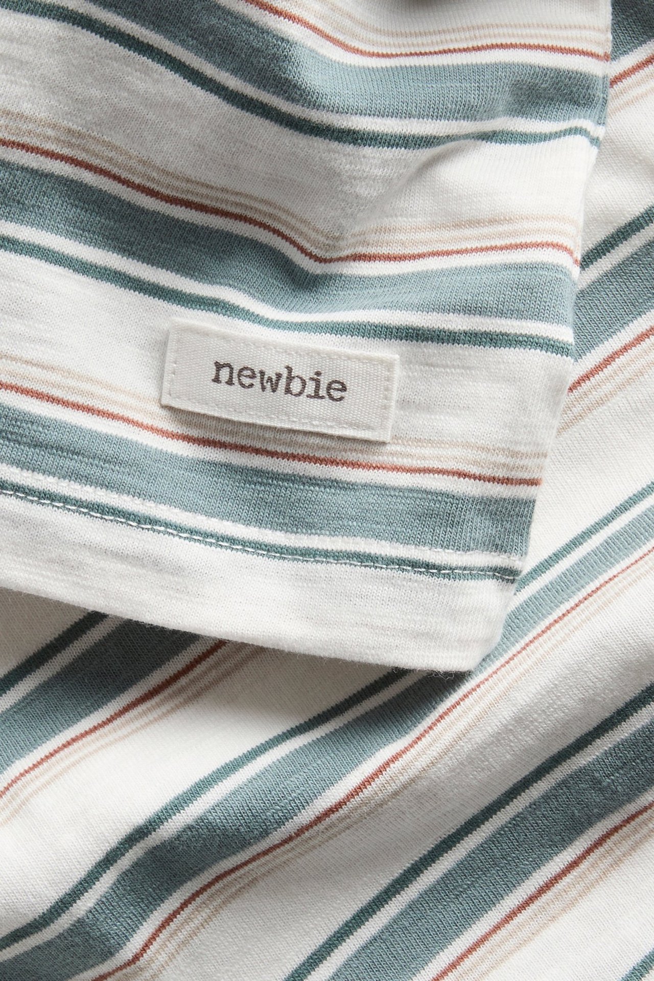 Nahaufnahme eines gestreiften, naturweißen Slub-T-Shirts mit grünblauen, braunen und beigen Streifen, Newbie-Etikett.