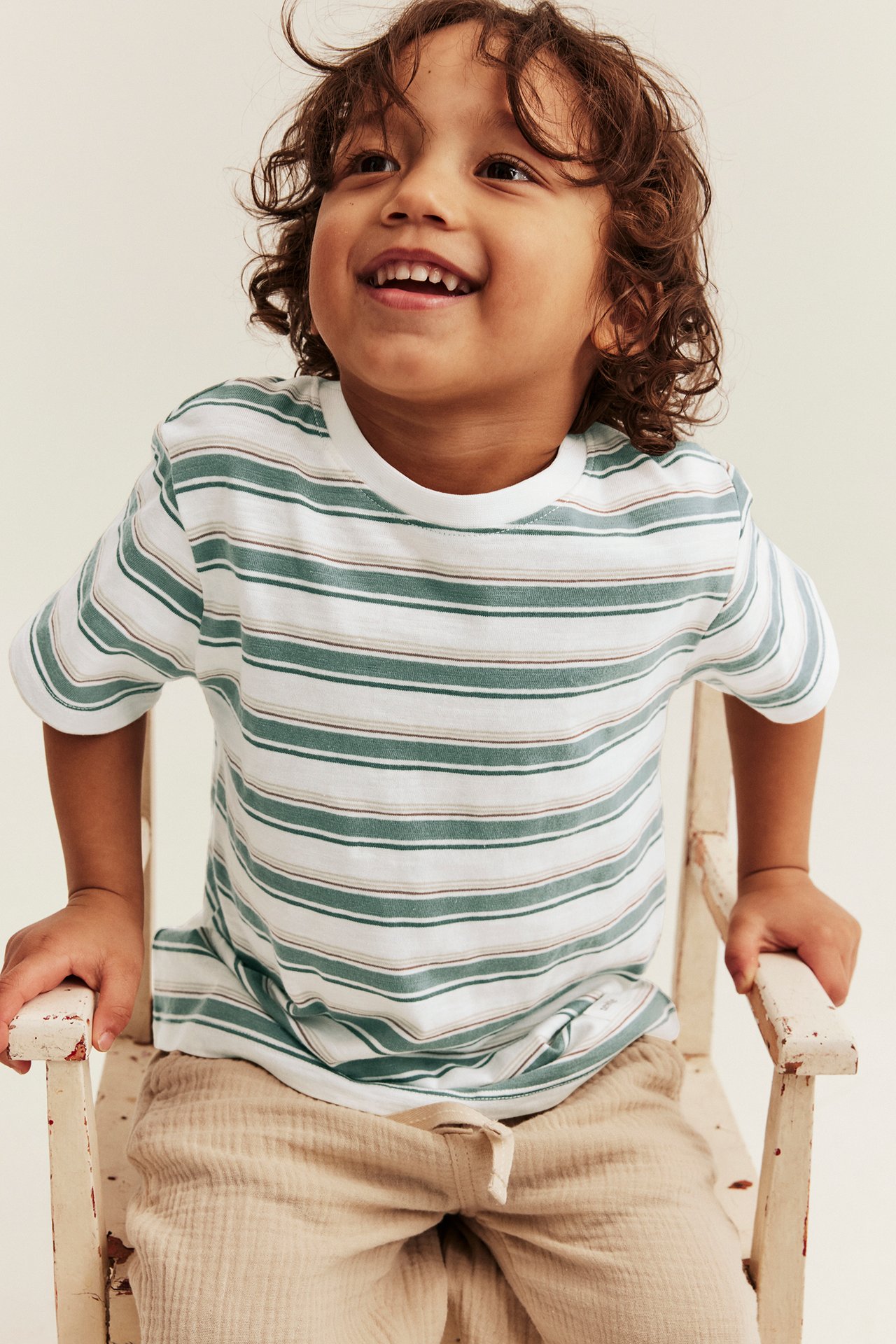 Front view: Kid in an off-white and green striped t-shirt with rolled cuffs and beige trousers.