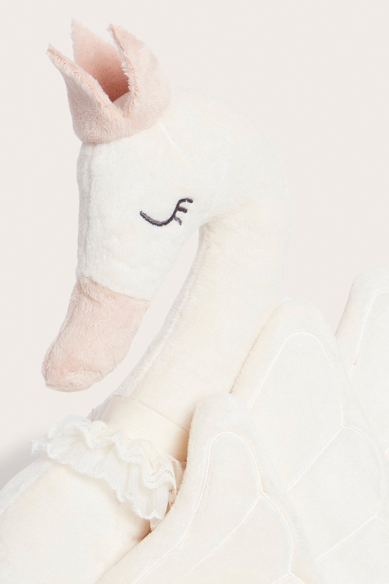 Front view of an off-white plush swan-shaped pillow with a light pink crown and beak, featuring ruffled trim.