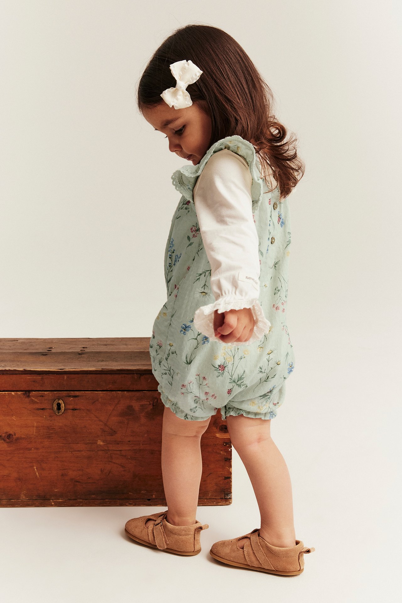 Side view of baby in light green floral cotton romper over white top, with white bow and brown shoes.