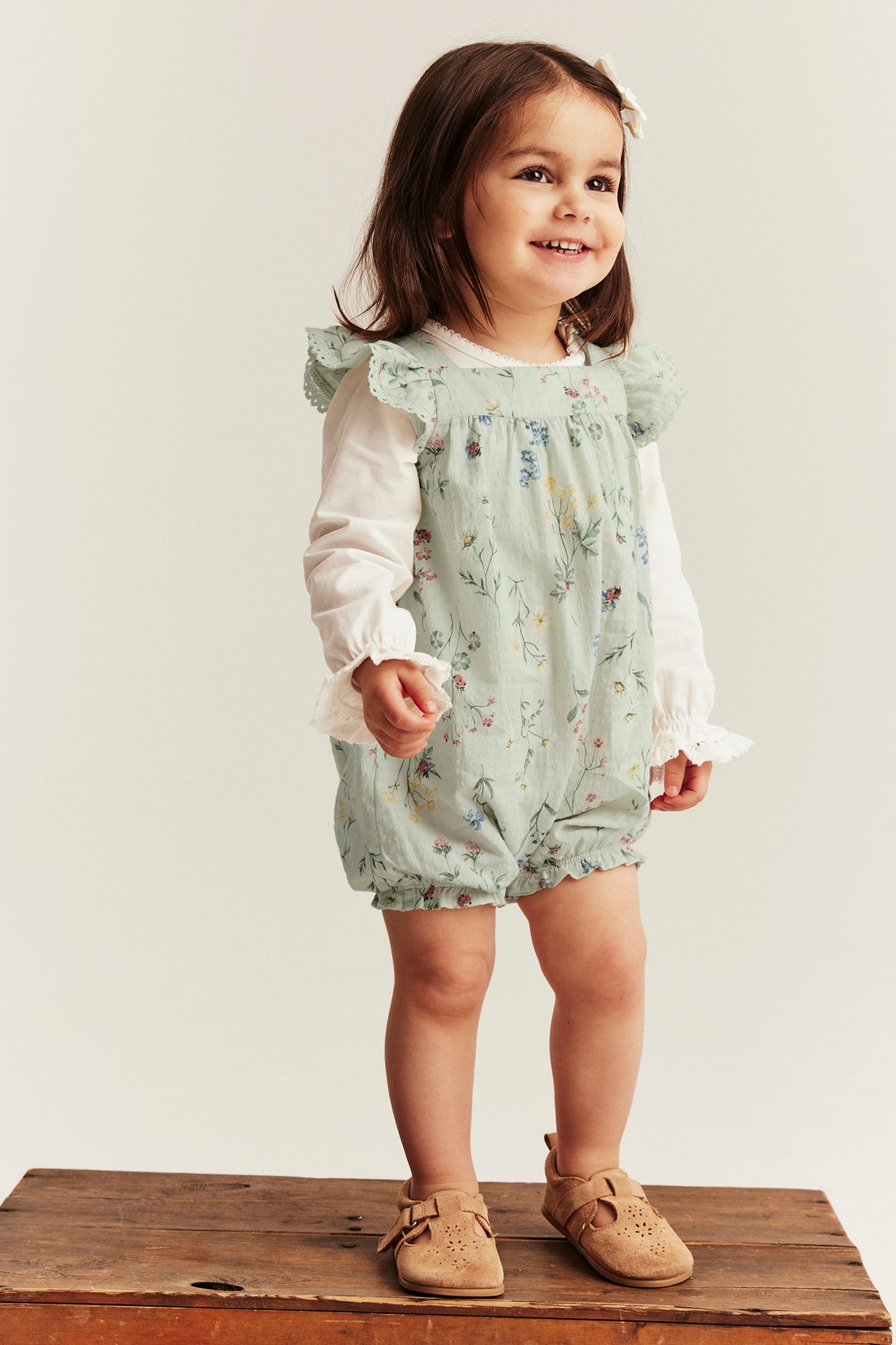 Vorderansicht: Baby-Mädchen in hellgrünem geblümtem Strampler, weißem Langarm-Top und braunen Wildlederschuhen.