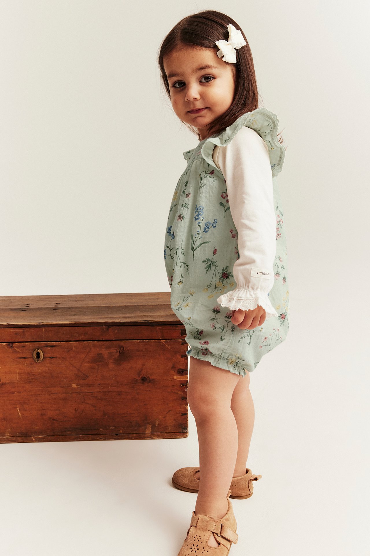 Front view: baby in a light green floral cotton romper, white long-sleeve top, brown shoes, white bow.