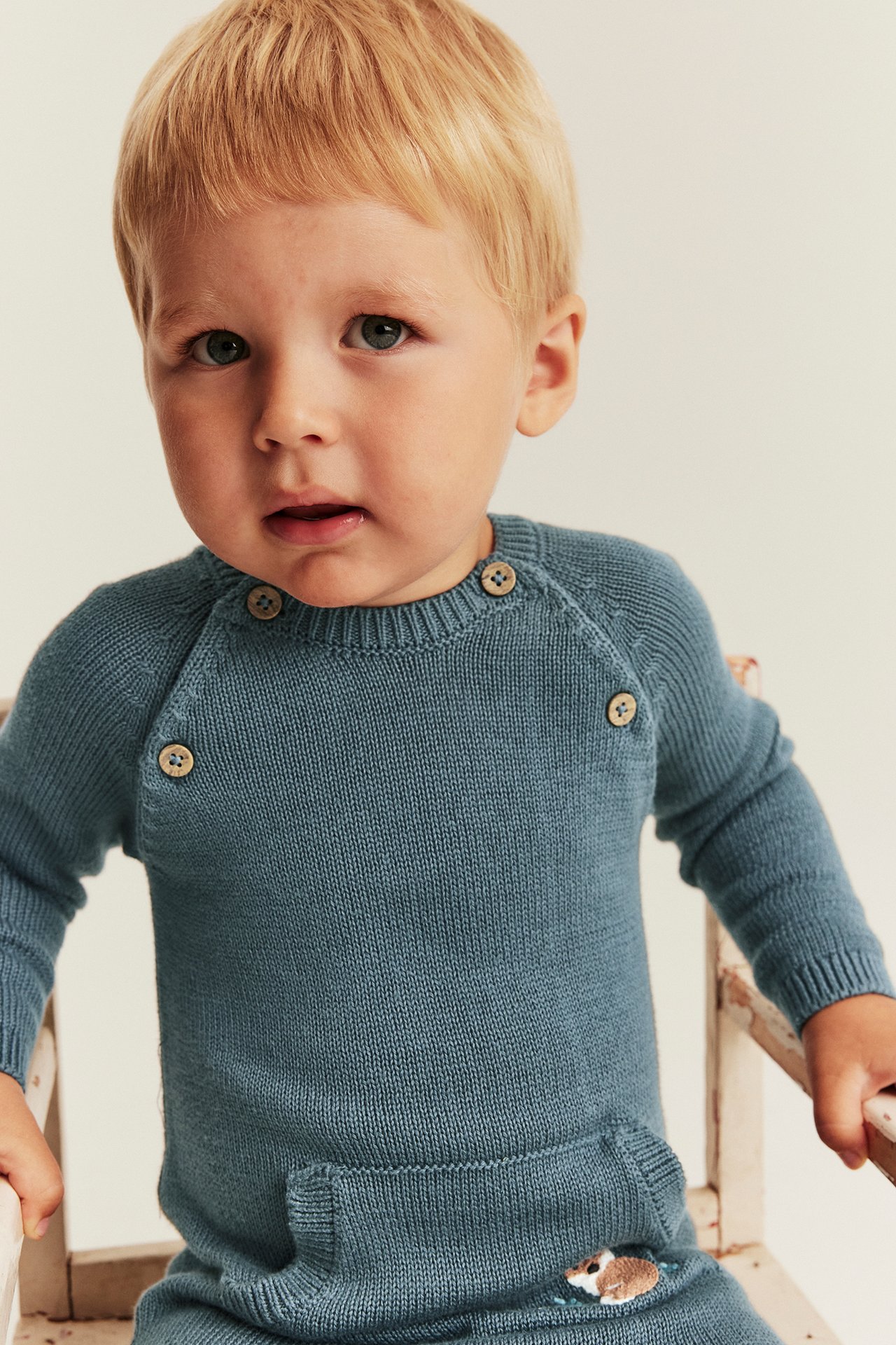 Front view of a baby boy wearing a blue knitted jumpsuit with wooden buttons and an embroidered animal.