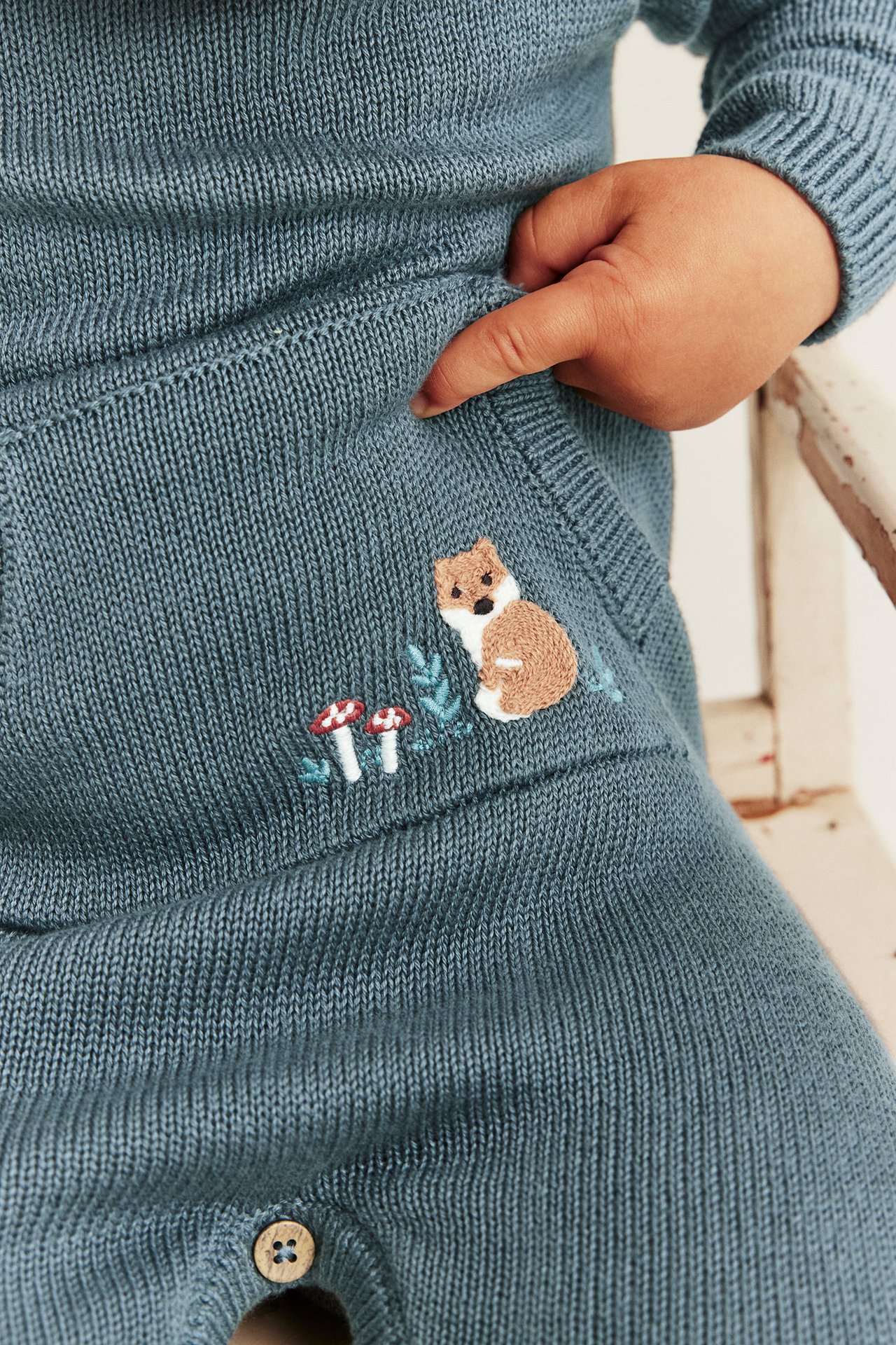 Close-up of a baby's teal knitted romper with embroidered fox and mushrooms.