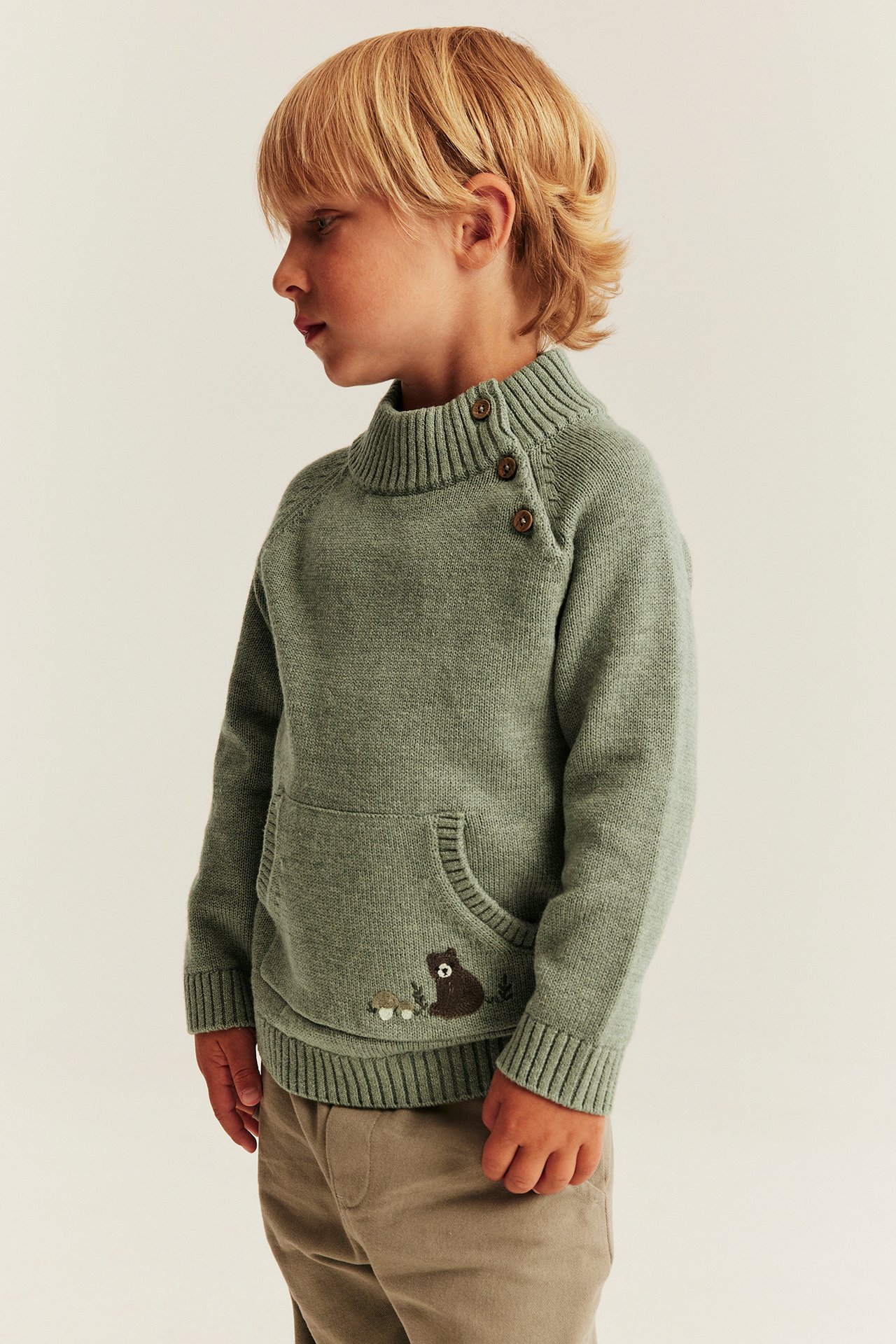 Seitenansicht eines Kindes in grünem Strickpullover mit Knöpfen am Kragen und Bärenstickerei.